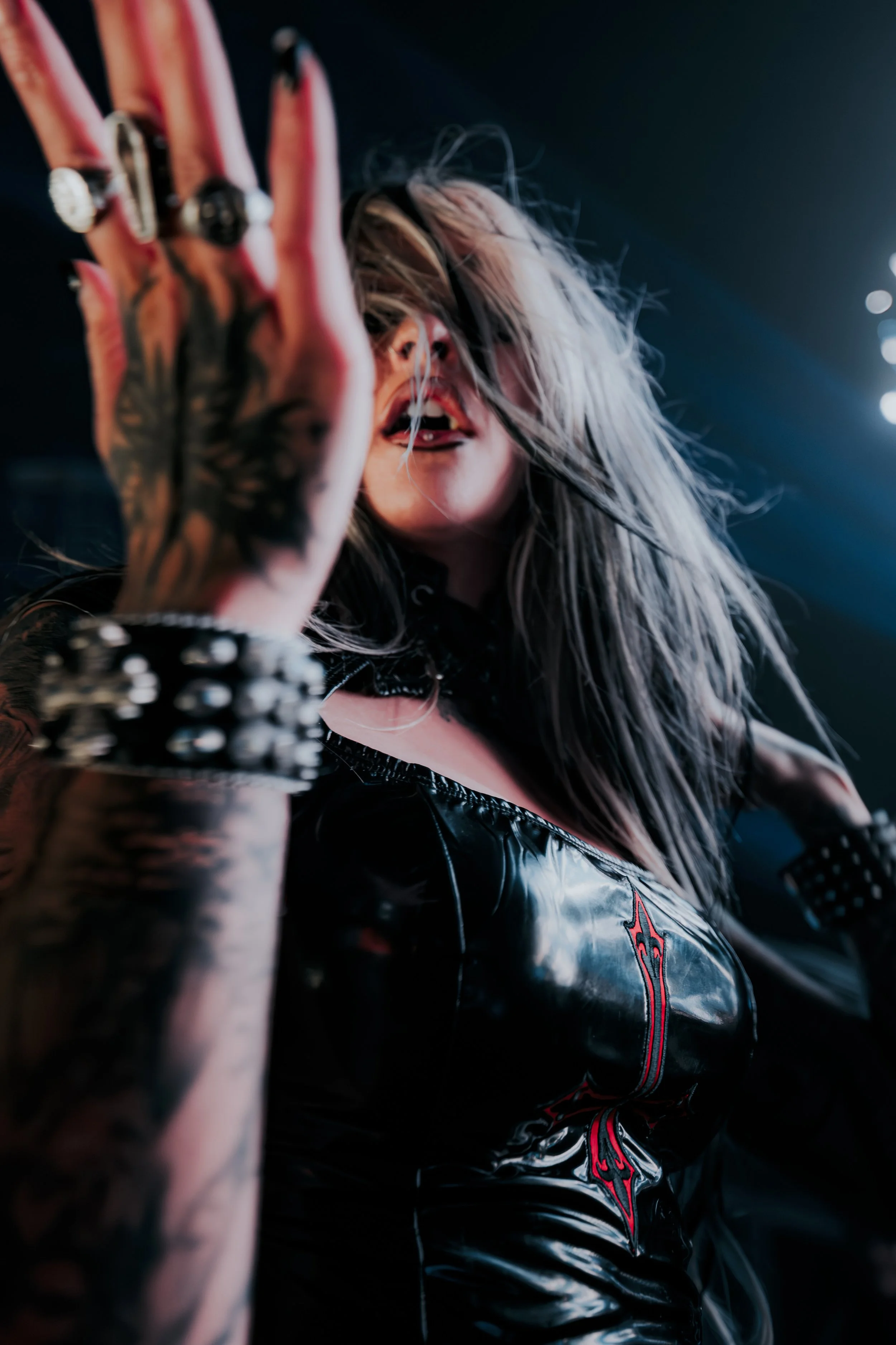 Close-up, low-angle shot of artist Luci in a black latex outfit with a red gothic cross. Her tattooed hand with silver rings and a spiked bracelet is in the foreground. Moody, cinematic concert lighting with a high-contrast aesthetic.