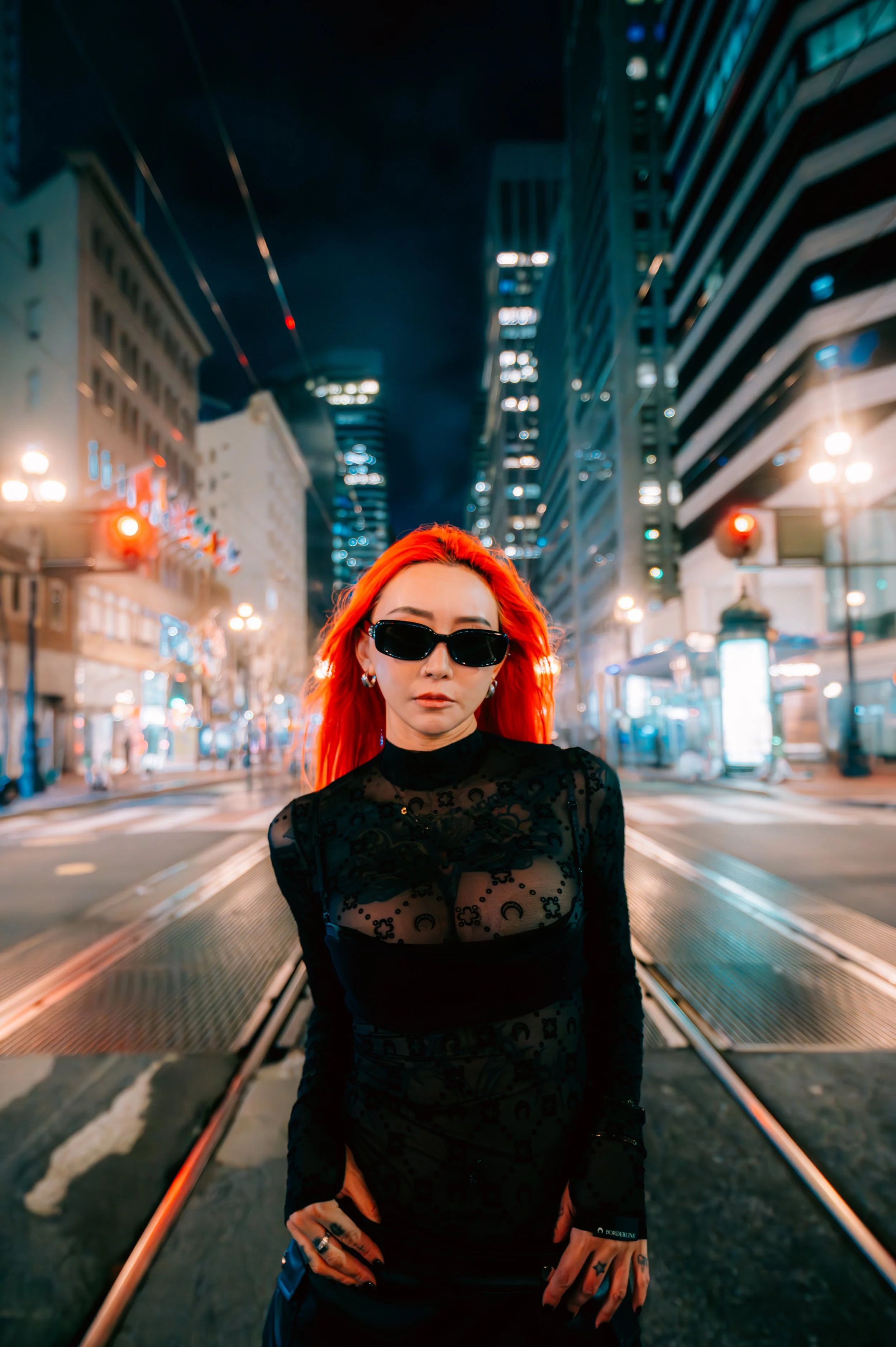Night portrait of artist Kromi standing on San Francisco city tracks. She has vibrant orange hair and wears black sunglasses with a sheer black top. The background features glowing city lights and skyscrapers with a cinematic urban feel.