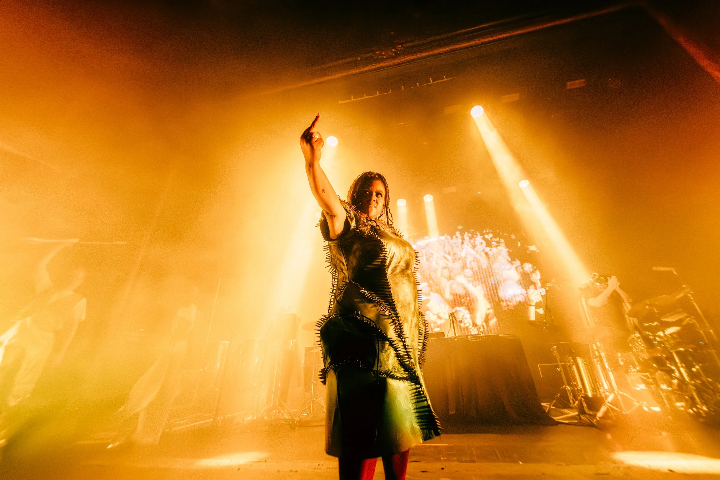 Wide-angle stage shot of a performer in a textured, spiky metallic dress raising her hand. Intense, golden-orange stage lights and thick atmospheric haze create a cinematic, high-energy glow. Powerful and moody concert photography.