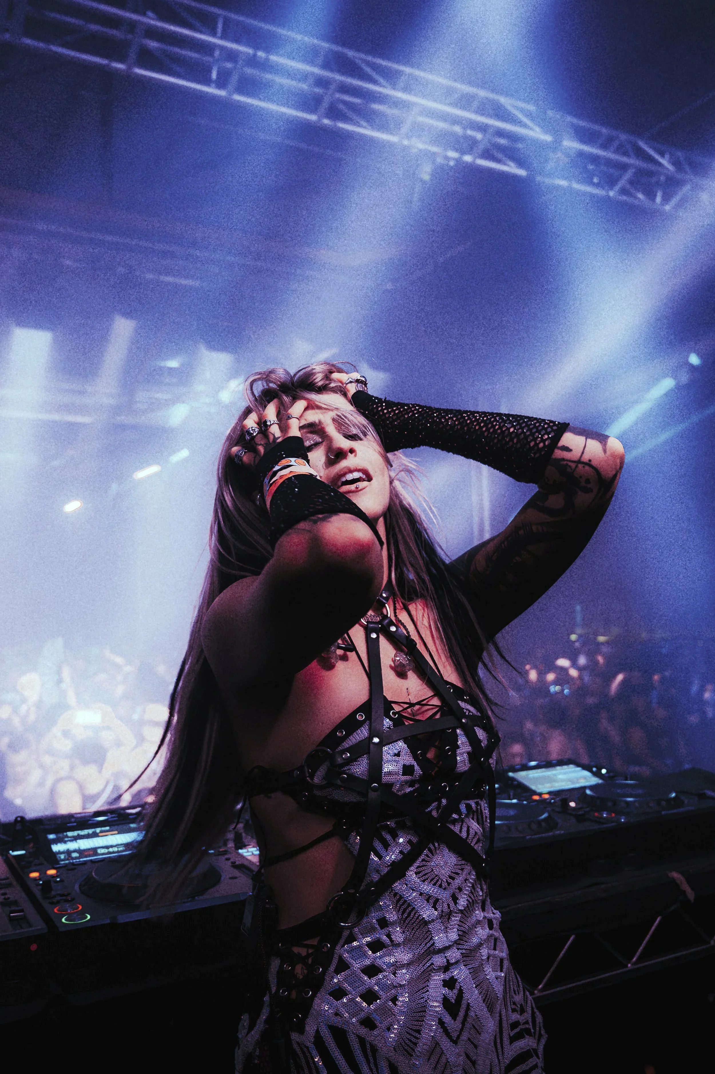 Candid shot of Luci performing at a DJ booth, hands in her hair with an expressive look. She wears a silver sequined outfit with a black harness and mesh sleeves. Vibrant blue stage lights and atmospheric haze create a cinematic festival feel.