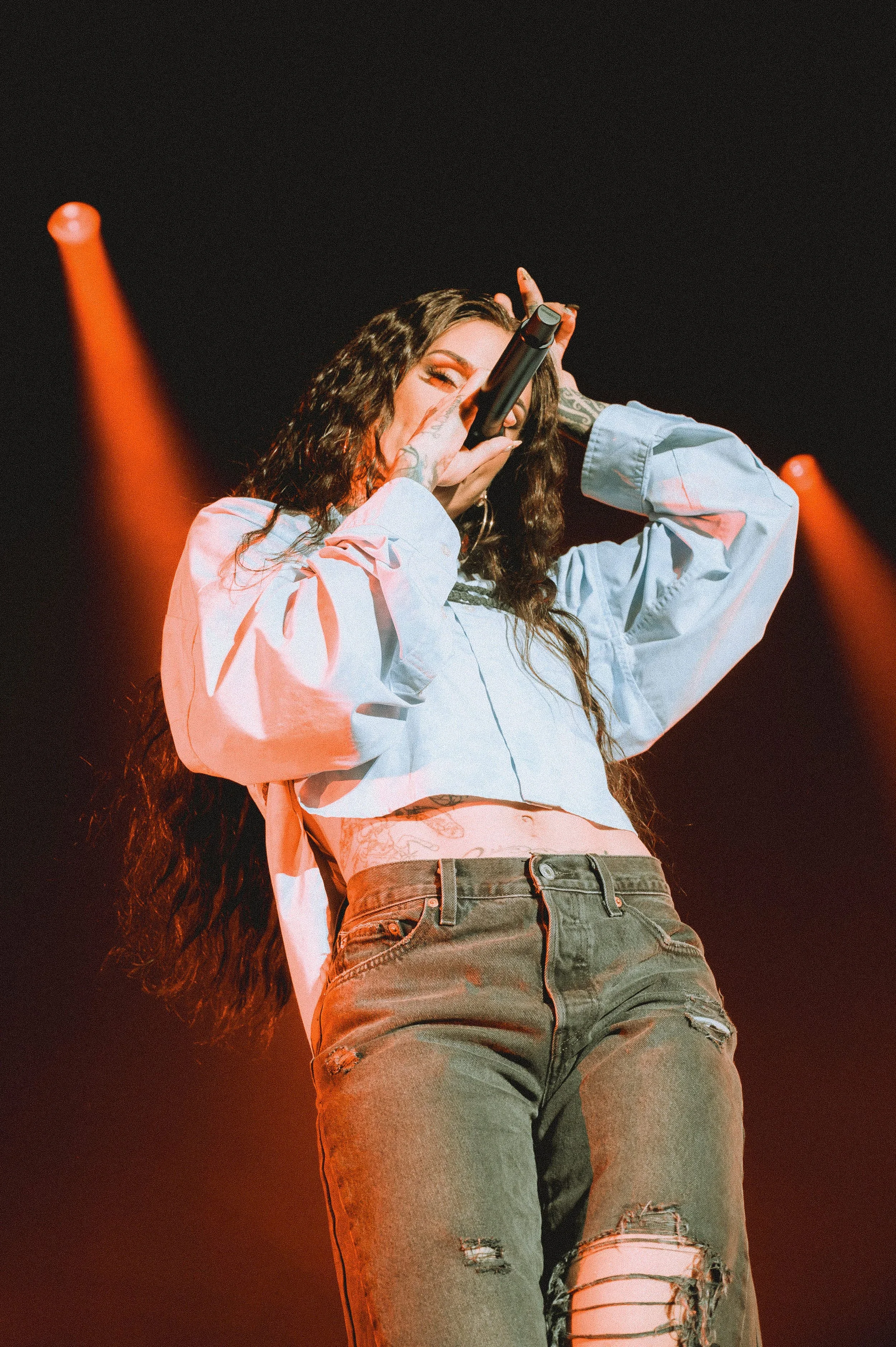 Low-angle concert photo of Kehlani singing into a microphone under warm orange spotlights. She wears a light blue cropped shirt and distressed denim jeans. The image features a heavy film grain texture and high-energy stage presence.