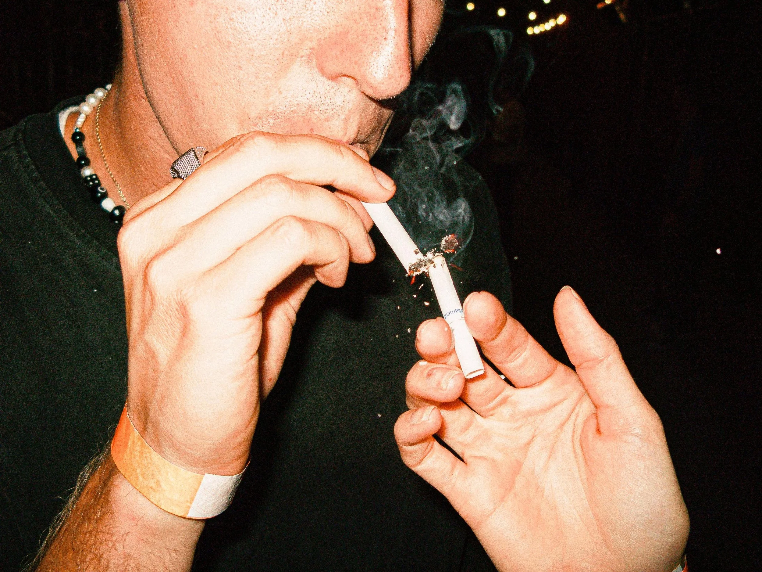 Close-up, high-flash photo of artist TOBEHONEST lighting a cigarette from another. Detailed shot of hands, jewelry, and smoke against a dark background. Raw, cinematic street-style photography with a heavy film grain aesthetic.