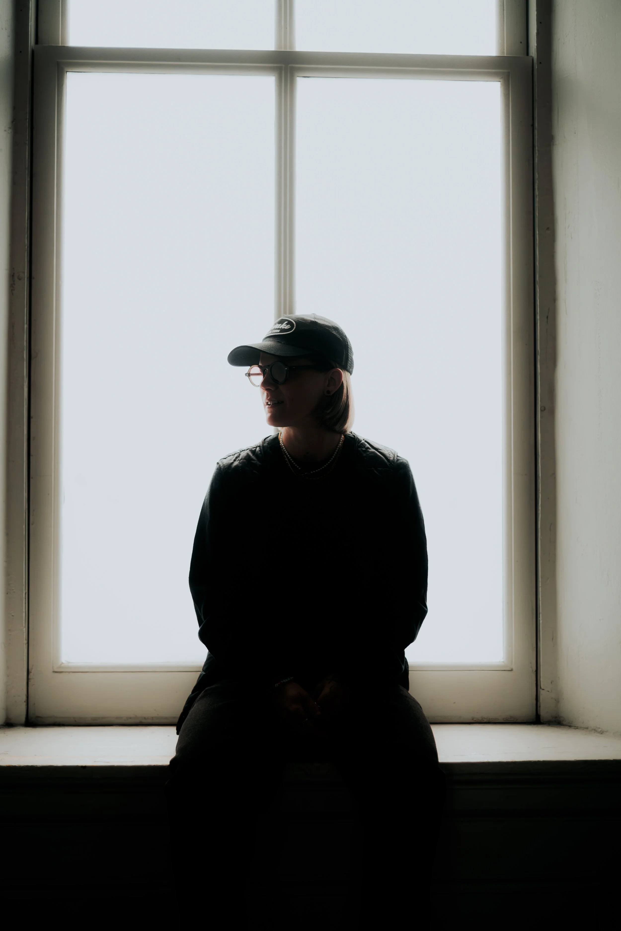 Silhouette portrait of artist J.Worra sitting on a windowsill against a bright, backlit window. She wears a baseball cap and glasses in a moody, high-contrast composition. Minimalist and cinematic aesthetic with a clean urban feel.
