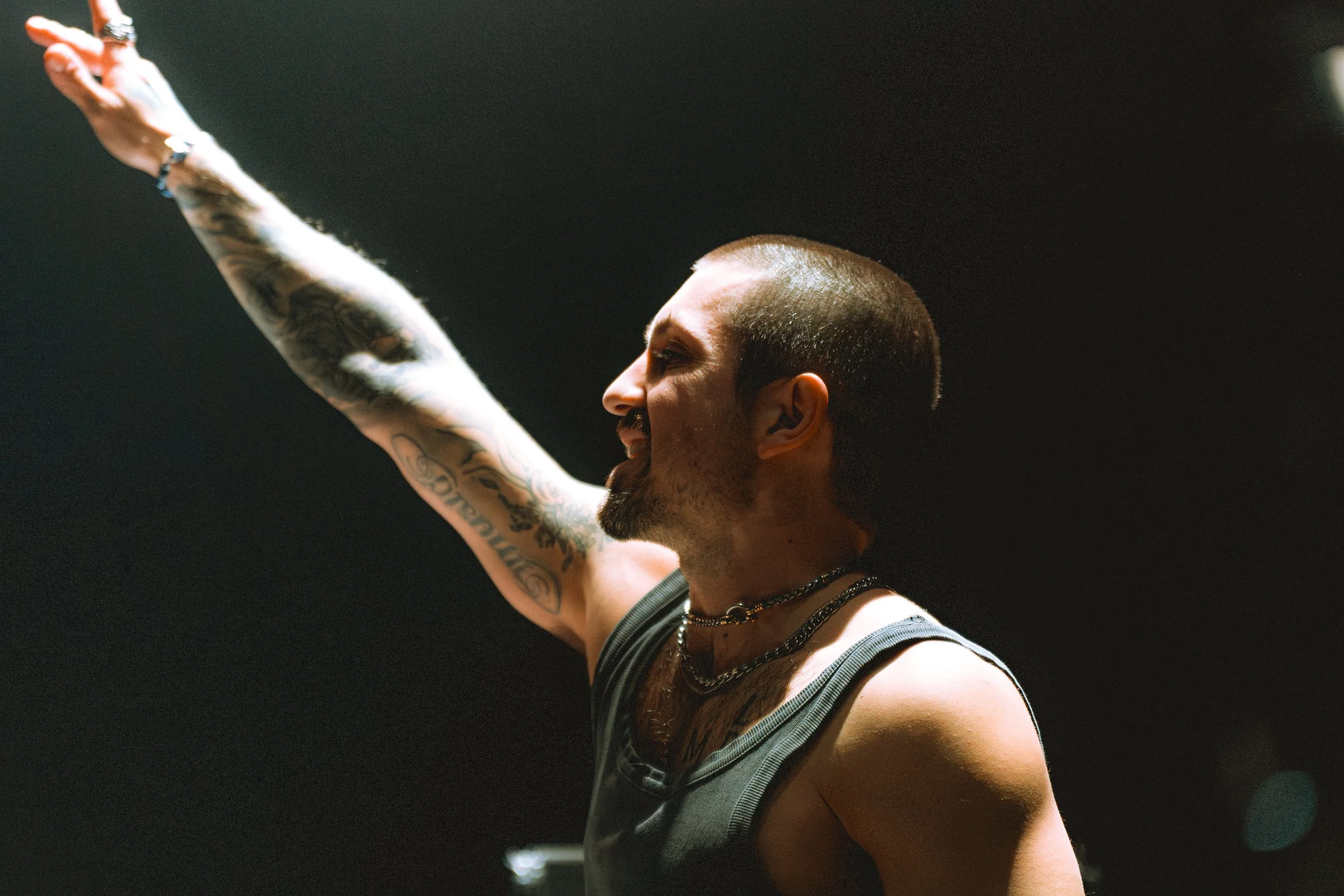 Side profile of artist Grabbitz performing on stage with his arm raised toward the light. He is wearing a dark tank top and necklaces, showing detailed arm tattoos. High-contrast concert lighting with a moody, dark background and film grain.