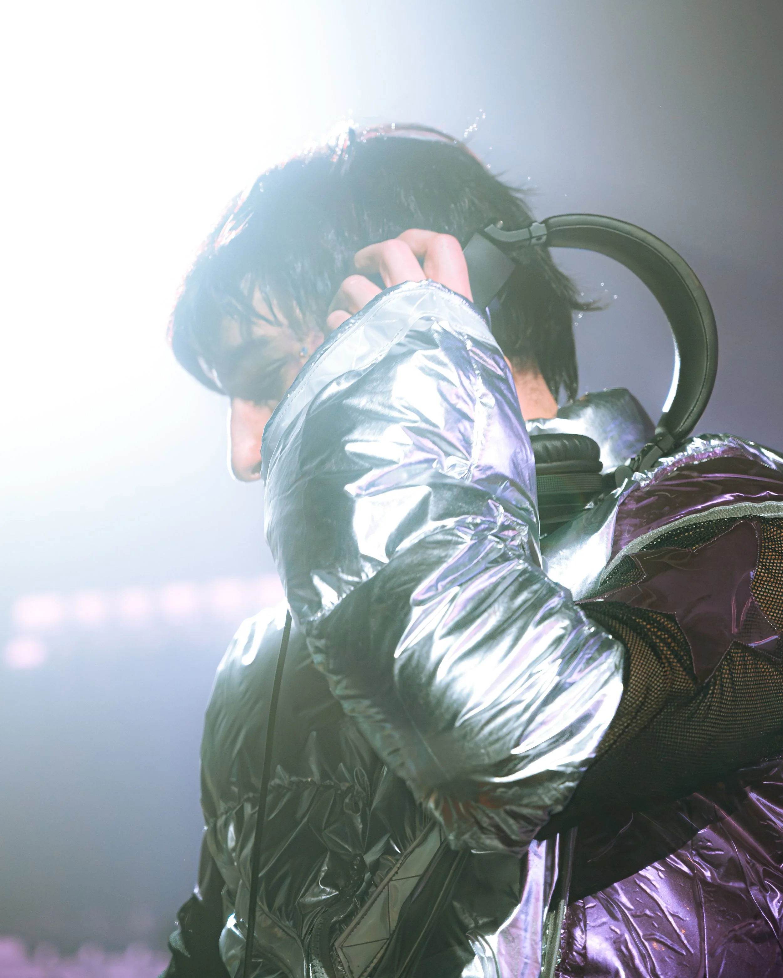 Close-up of Starjunk95 at Countdown NYE wearing a reflective silver and purple metallic jacket. He is adjusting his headphones under a bright, hazy stage light. Cinematic festival shot with high-contrast highlights and a futuristic aesthetic.