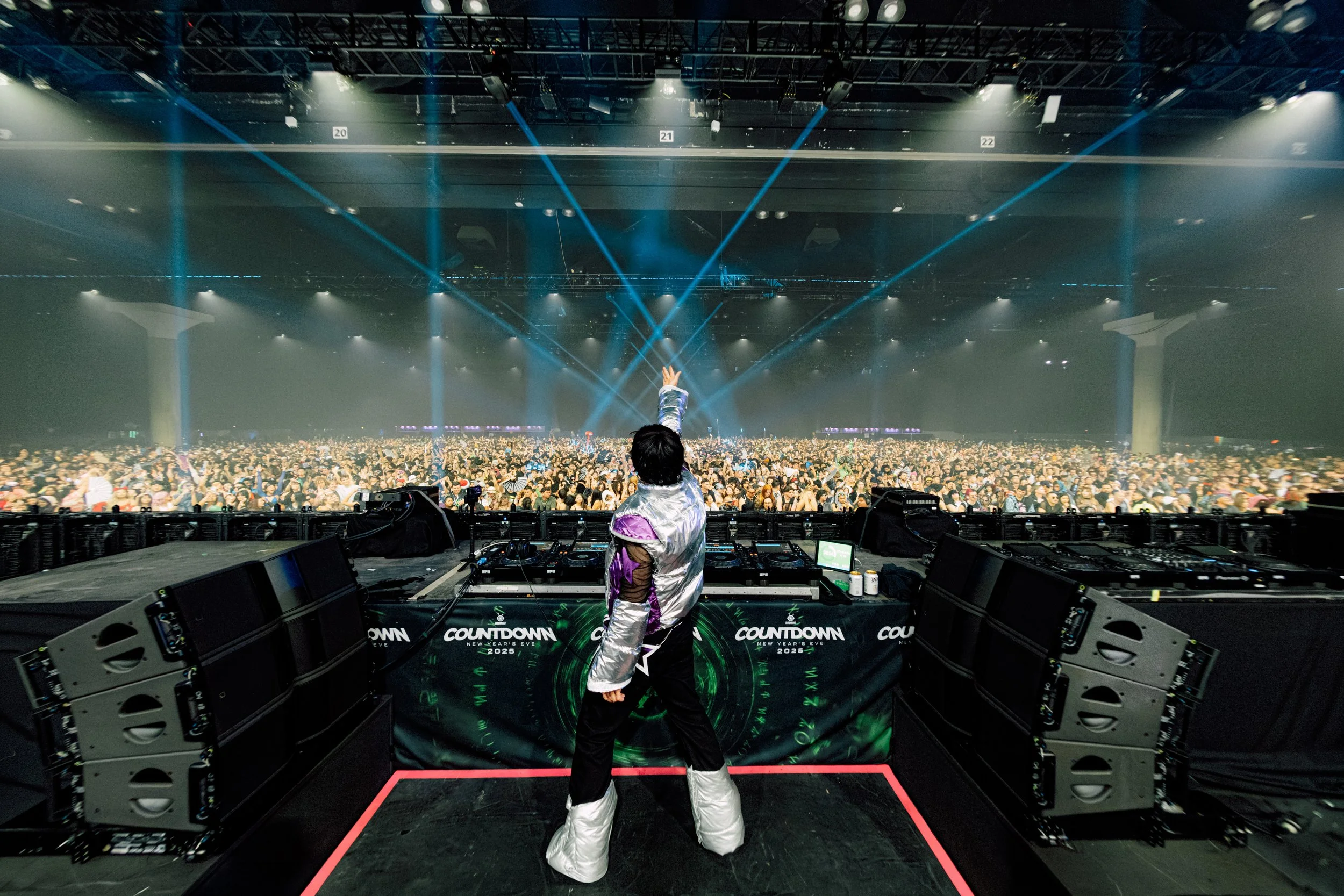Wide-angle shot from behind Starjunk95 at Countdown NYE. Wearing a metallic silver and purple outfit, he points toward a massive crowd under blue lasers. The "Countdown" branded stage and high-energy festival atmosphere are in full view.