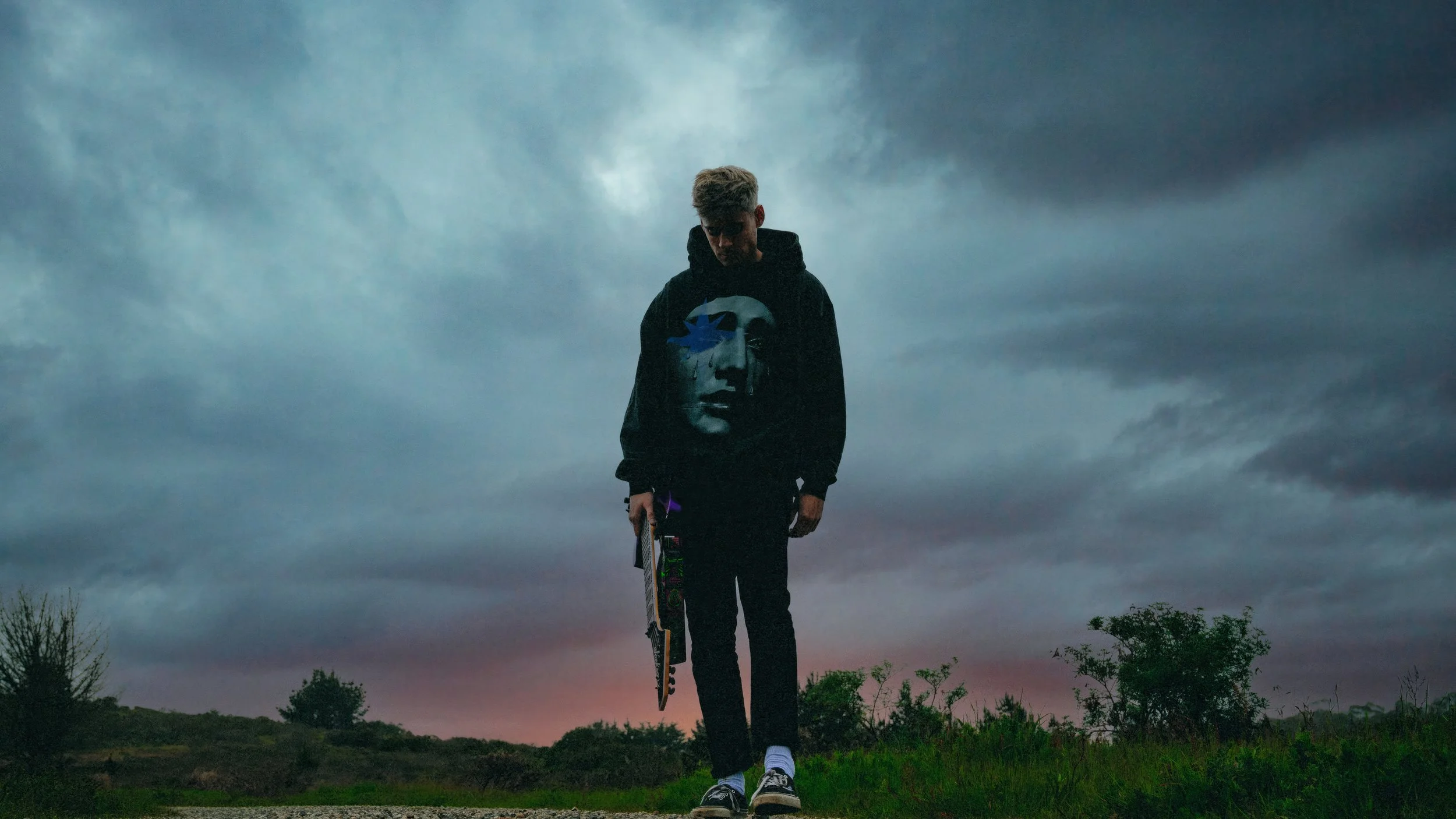 Wide-angle outdoor portrait of artist Fairlane standing under a dark, moody storm sky. He is wearing a black hoodie with a large face graphic and holding a guitar. Cinematic, high-contrast edit with a dramatic and melancholic atmosphere.