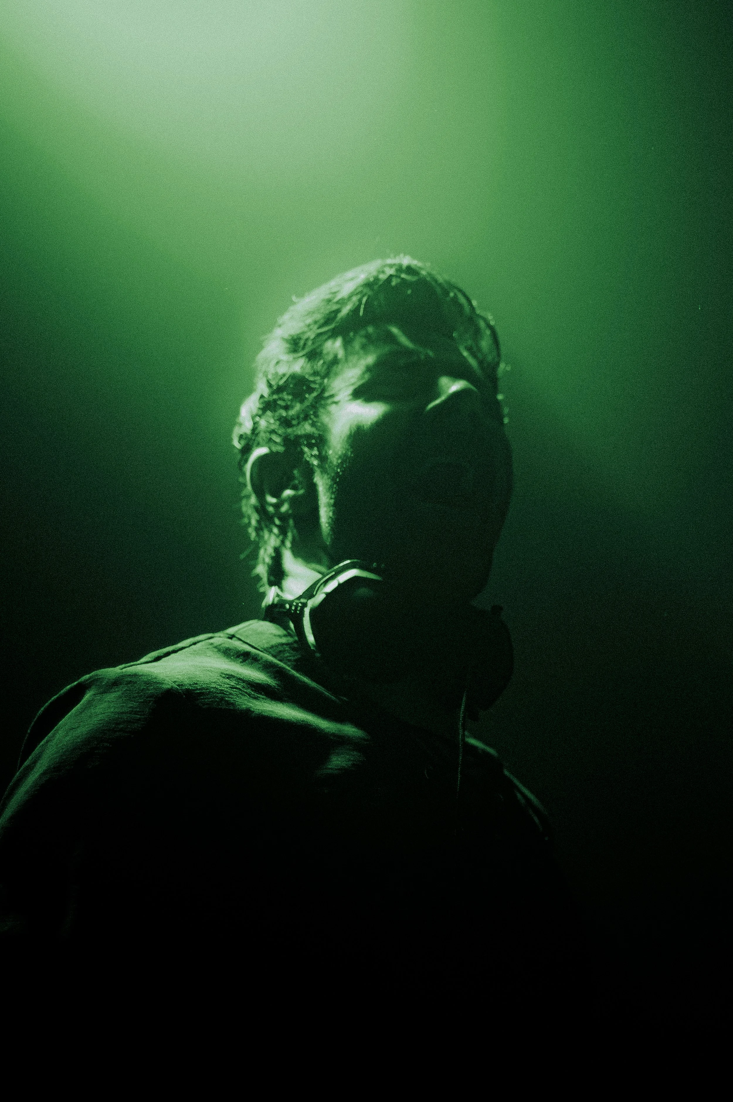 Moody, high-contrast portrait of artist KSHMR on stage, silhouetted against a vibrant green spotlight. He has headphones around his neck, and the image features a heavy film grain texture for a cinematic, underground concert feel.