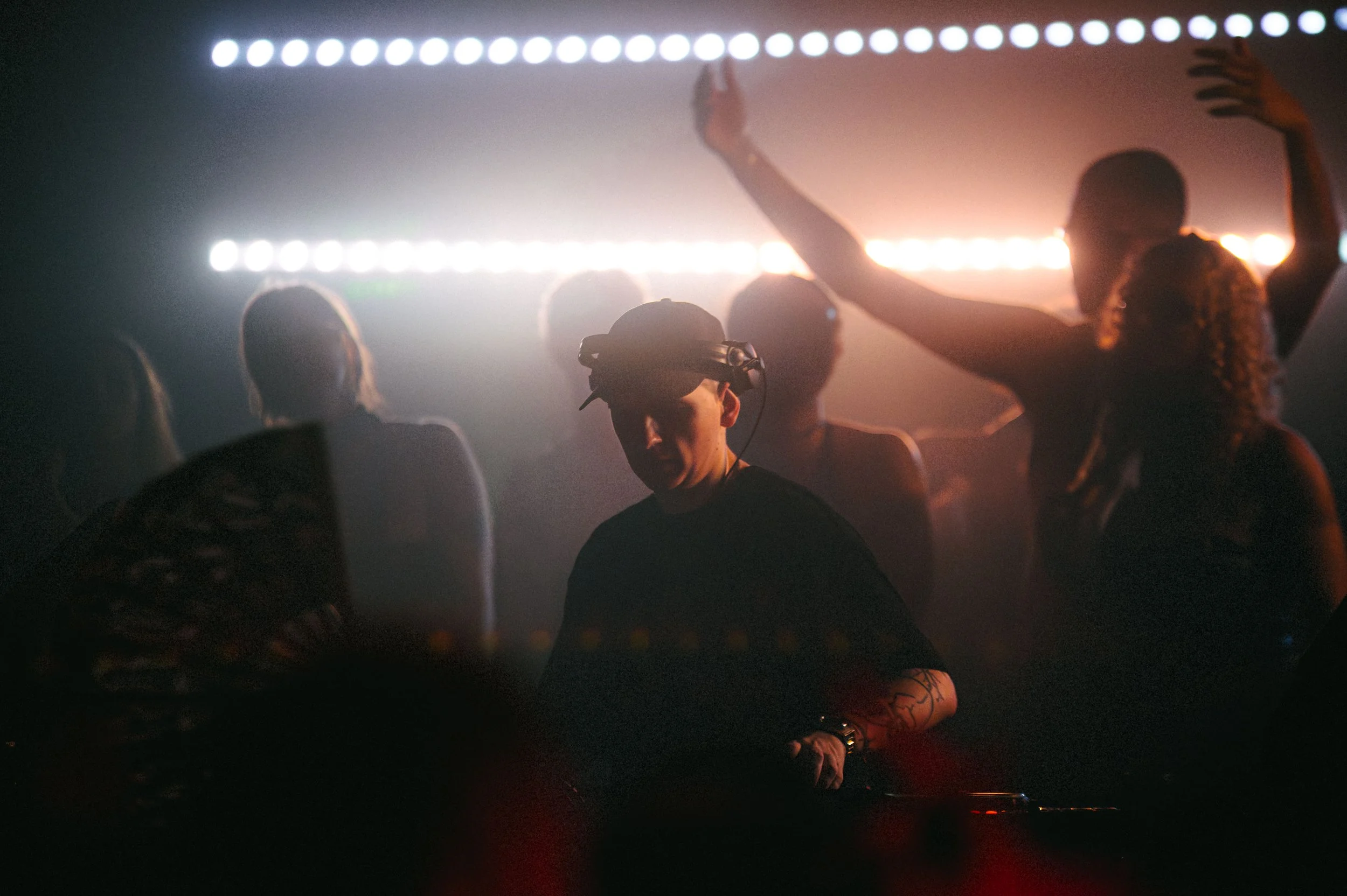 Candid shot of DJ klankluster performing behind the decks in a dark, atmospheric venue. Rows of bright white lights and a warm orange glow silhouette the crowd in the background. High-contrast concert photography with a cinematic, grainy feel.