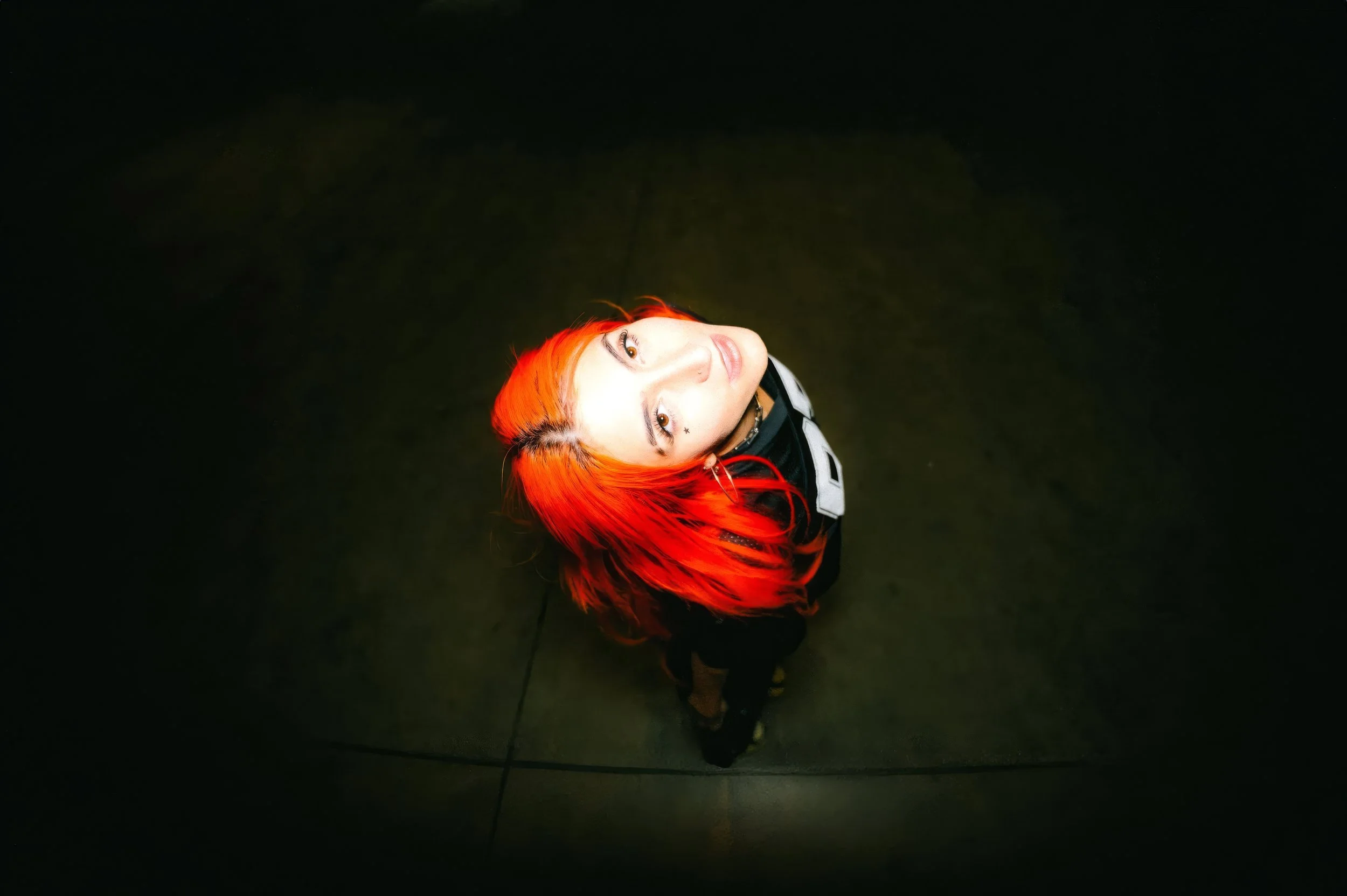 Overhead fisheye lens portrait of artist Nostalgix with vibrant, fiery orange hair. She looks directly up at the camera against a dark, moody concrete floor. Cinematic wide-angle effect with heavy vignetting and high-contrast lighting.