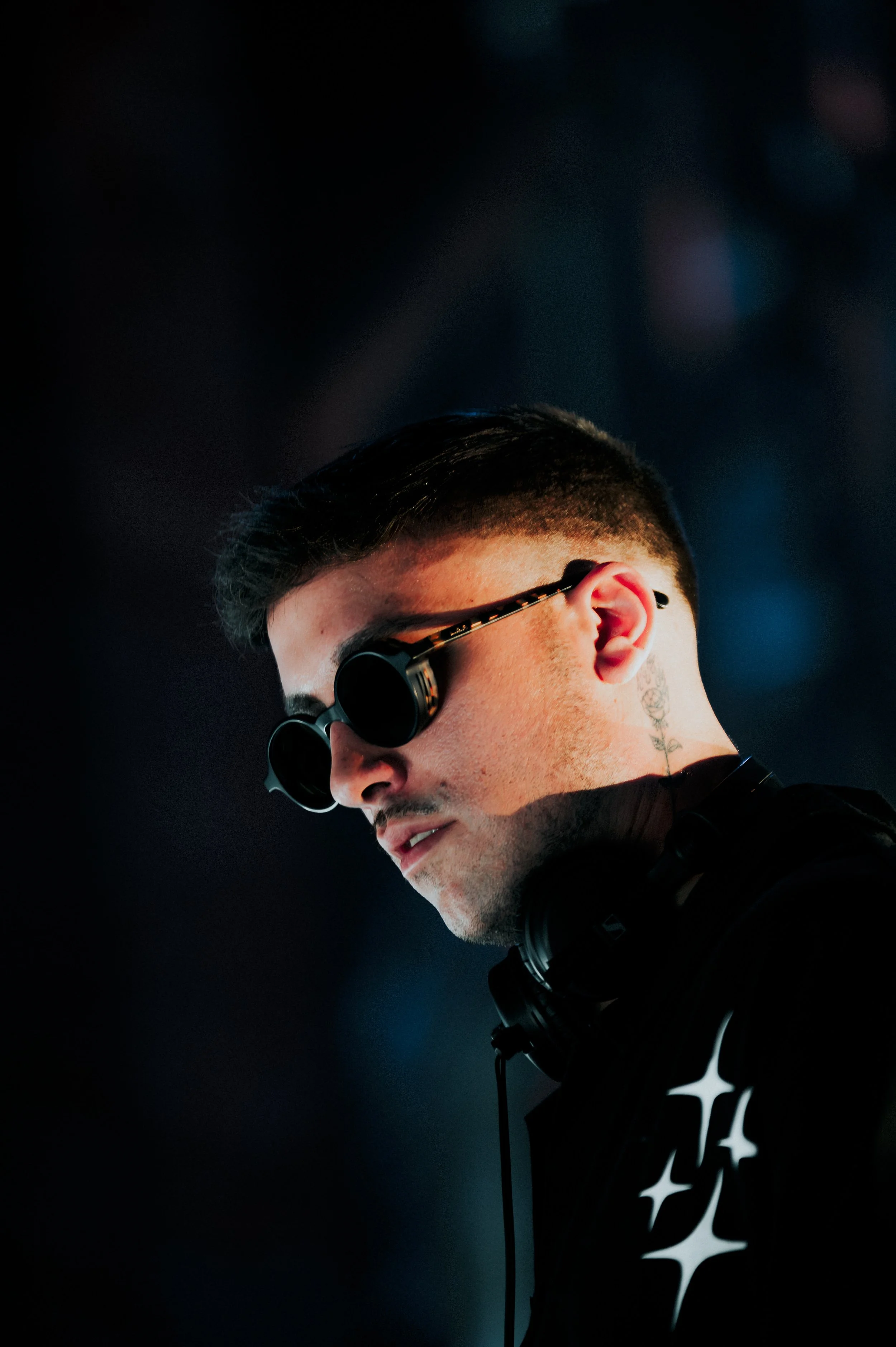 Close-up side profile of Matroda at EDC LV, wearing round sunglasses and headphones. The shot features high-contrast lighting, a subtle rose neck tattoo, and a black jacket with star graphics. Cinematic, moody festival photography.