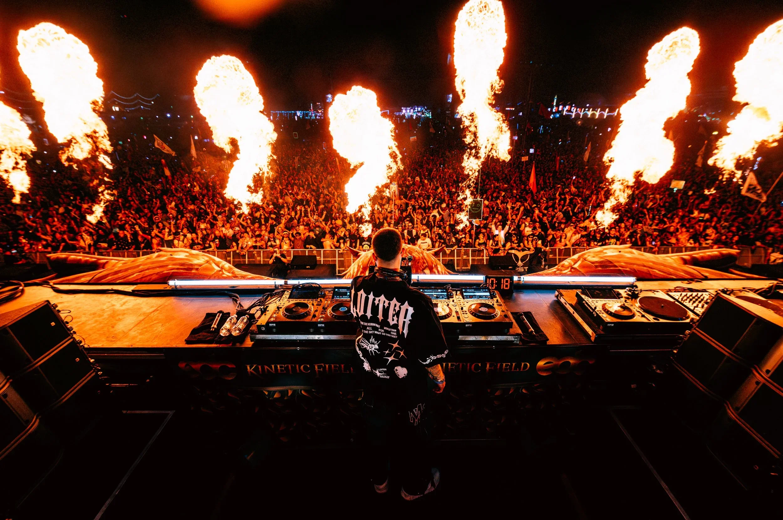 Over-the-shoulder shot of Matroda performing at the EDC Kinetic Field stage. Massive fire cannons erupt behind the DJ booth, illuminating a sea of thousands of fans. Intense, high-energy festival atmosphere with warm orange pyrotechnics.