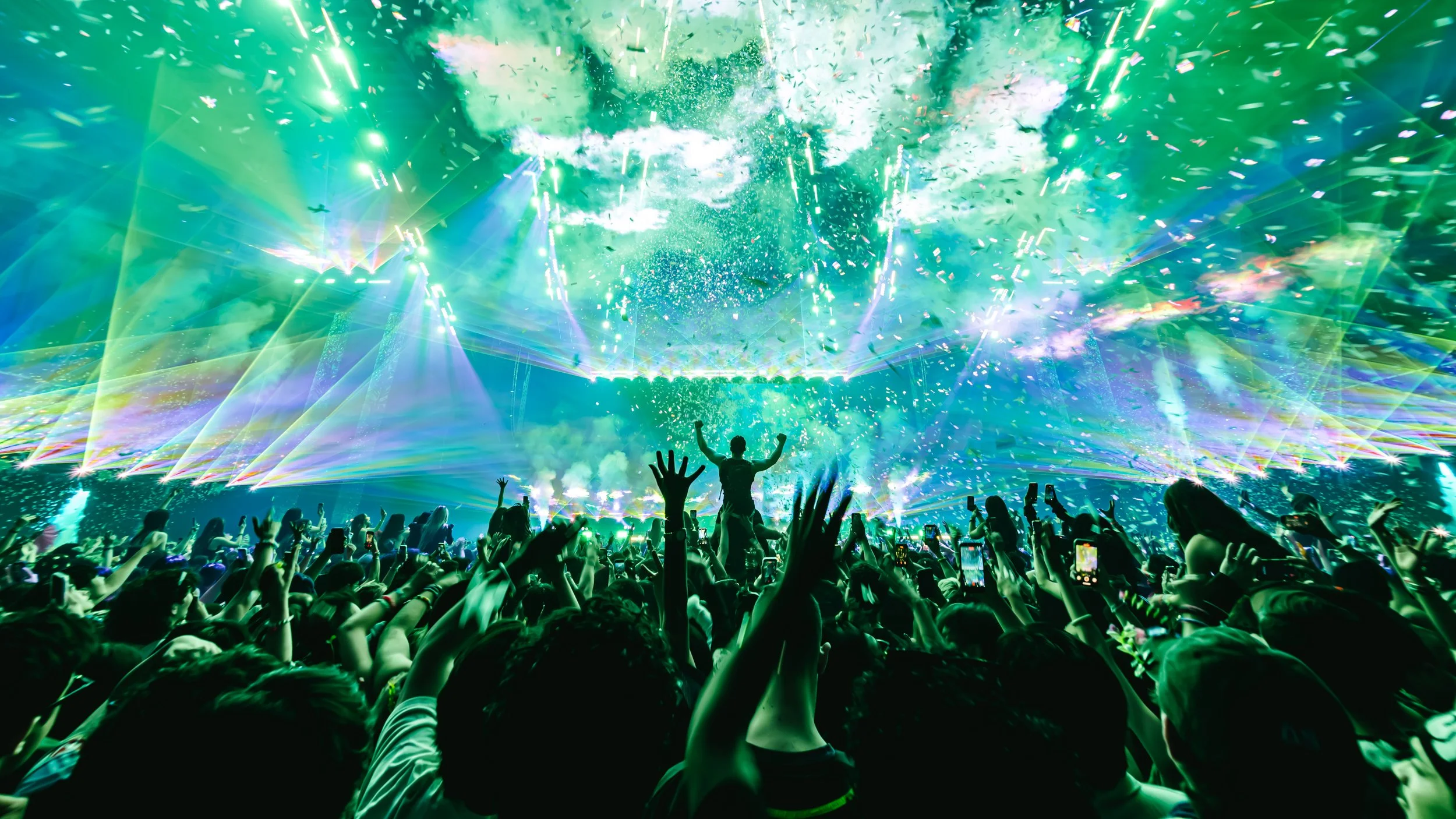 Wide-angle shot of a massive crowd at an Excision show with vibrant green and blue lasers filling the air. Confetti falls over fans with their hands raised in a high-energy atmosphere. Dramatic, cinematic festival production at its peak.