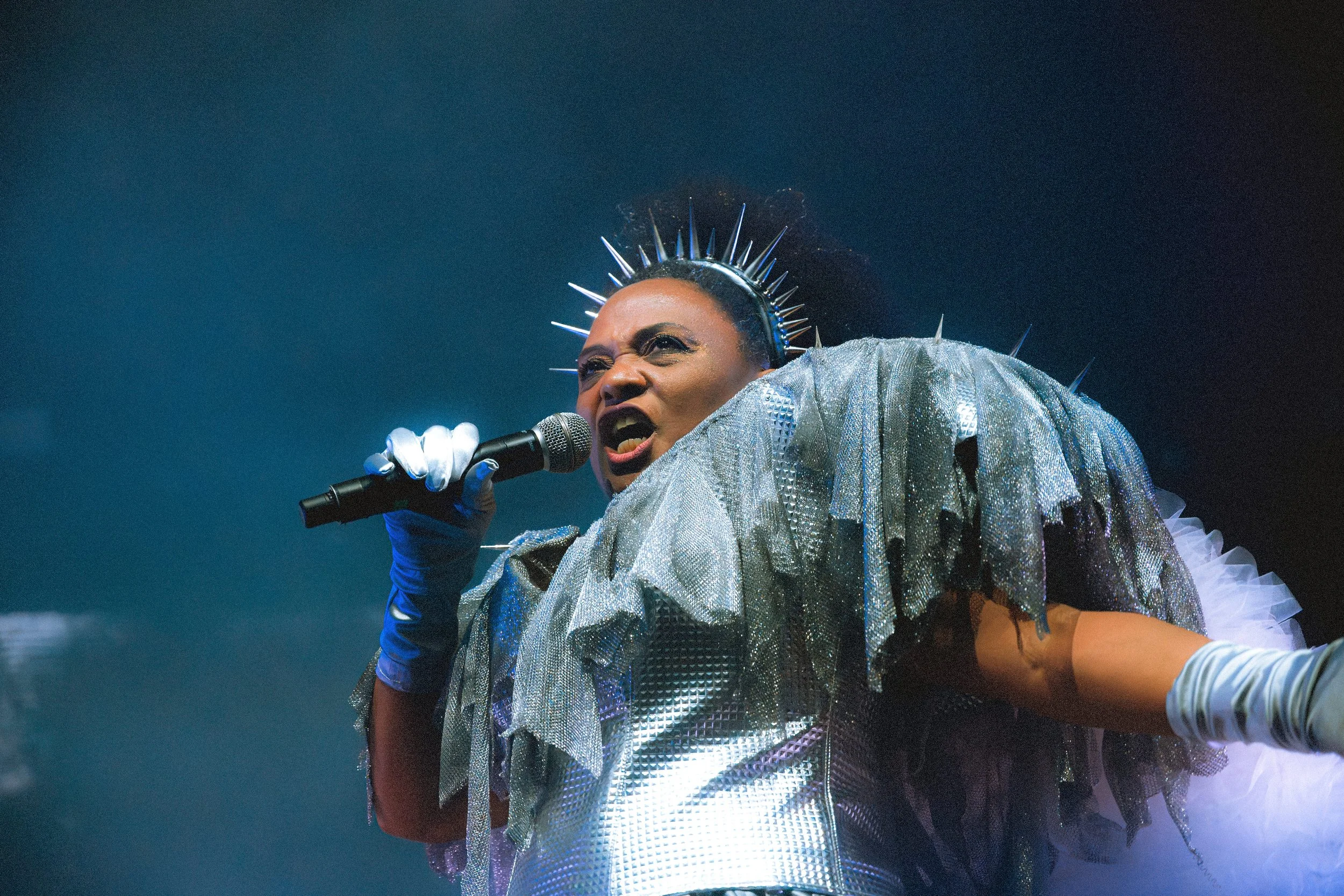 Live performance of a singer with a spiked crown and shimmering silver fringe outfit. She is singing into a microphone with high energy against a dark blue background. The photo features cinematic stage lighting and film grain.