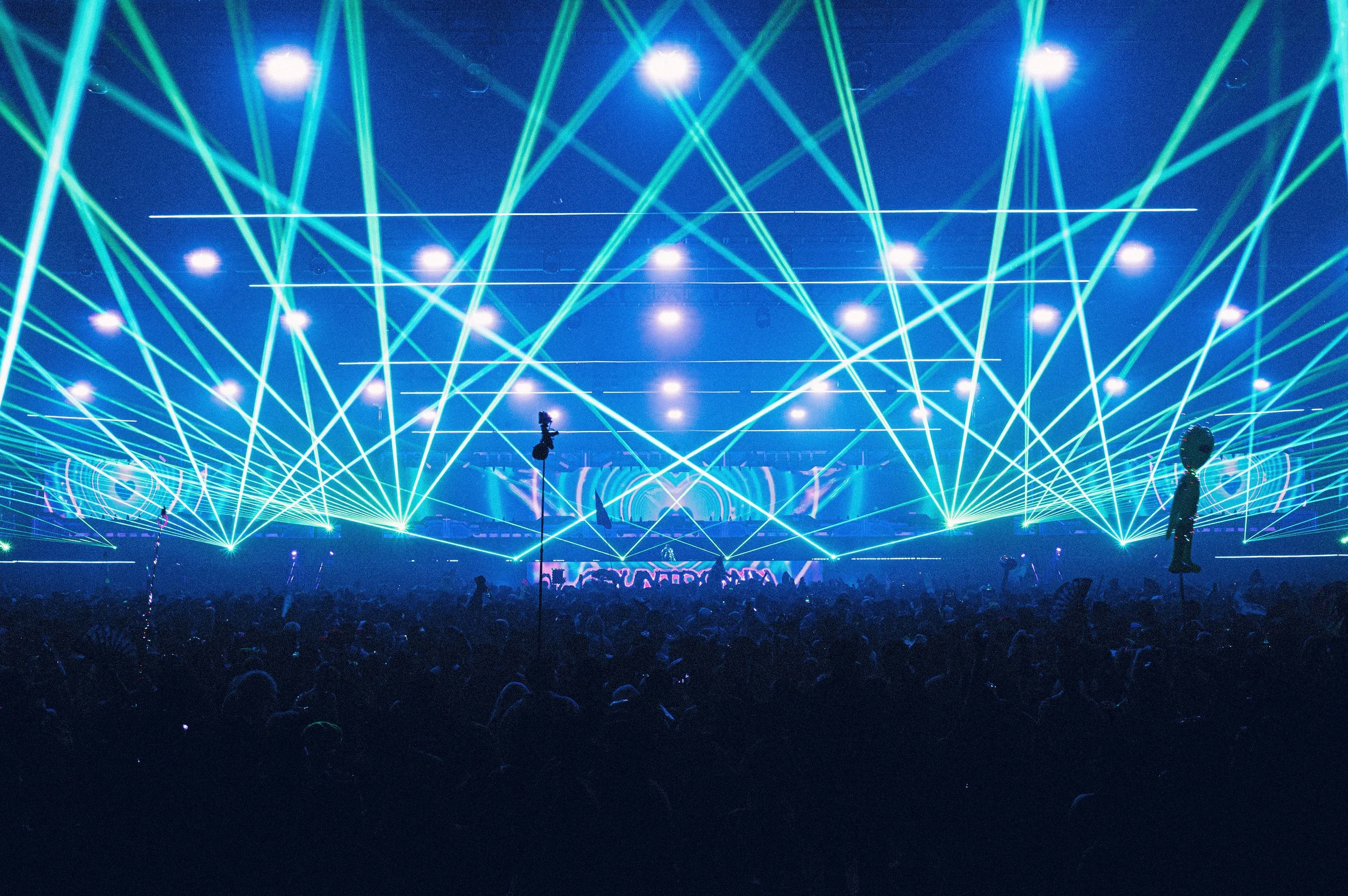 Vibrant wide-angle shot of Starjunk95 performing at Countdown NYE. Electric blue and green lasers crisscross over a massive crowd. High-energy festival production with bright stage lights and a cinematic, grainy concert photography style.