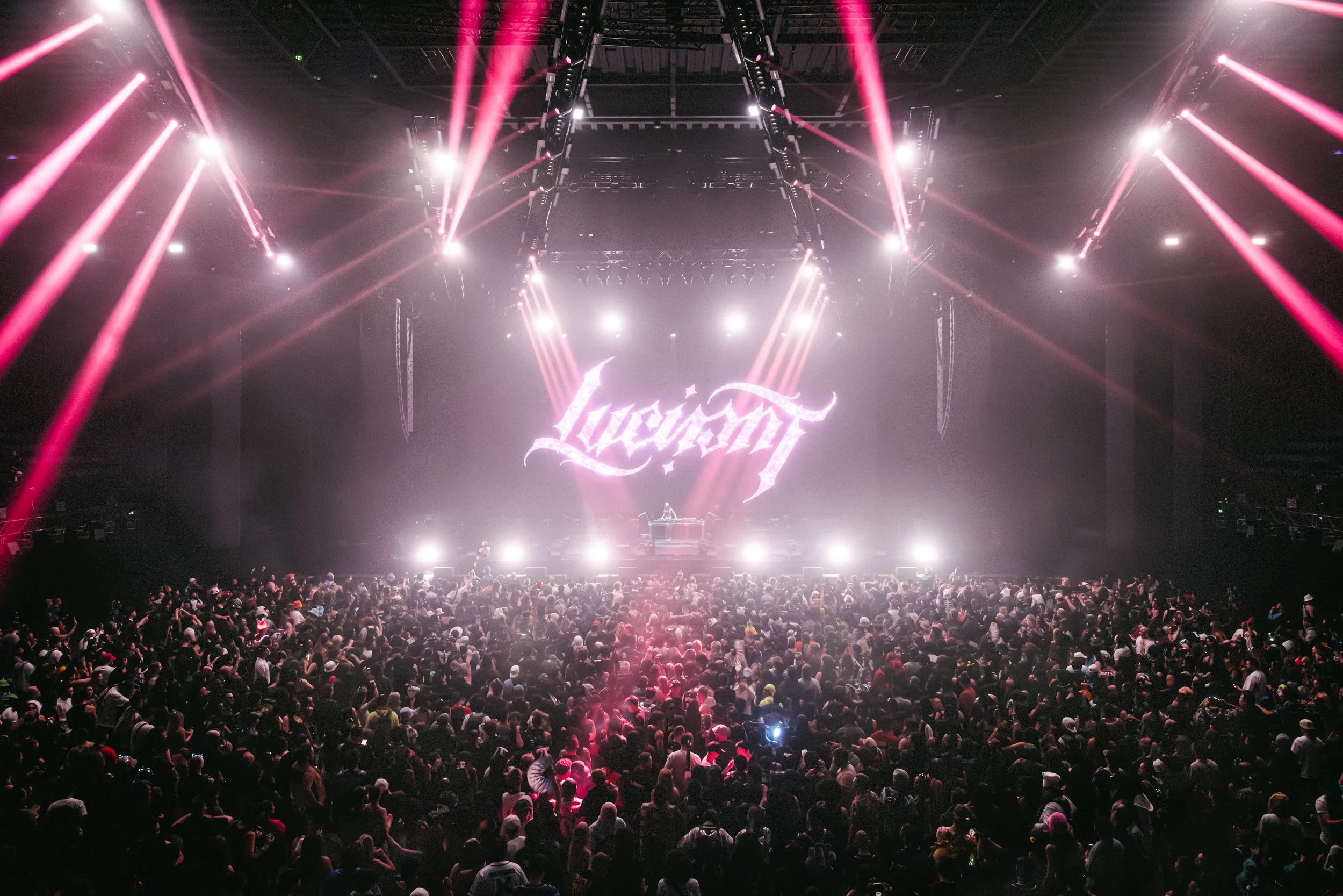 Wide-angle shot of artist Luci performing for a massive indoor crowd. Bright pink beams of light radiate from the stage, where her logo is displayed on a large screen. High-energy festival atmosphere with dramatic, cinematic lighting.
