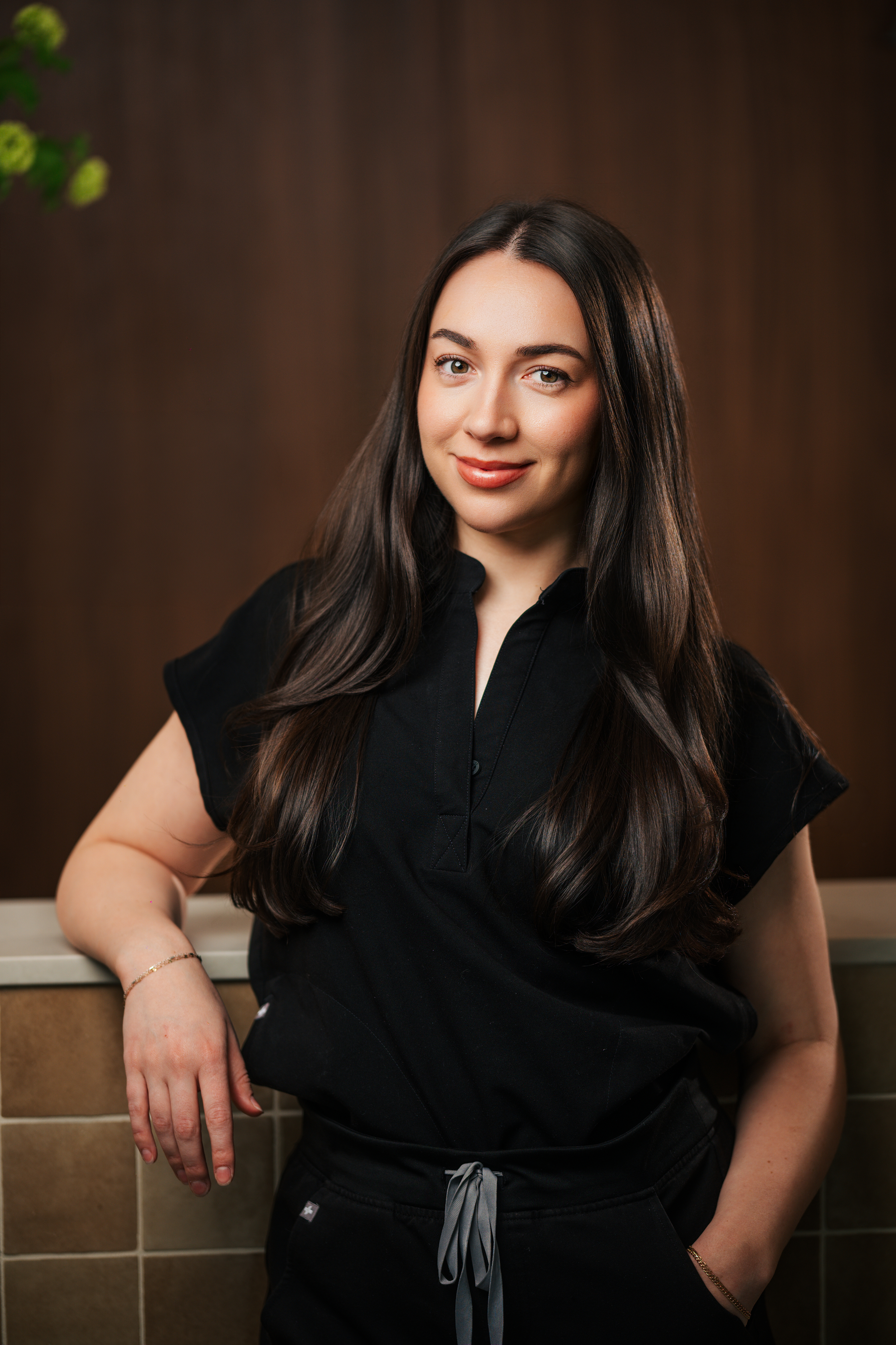 A young woman with long dark hair, wearing a black top and pants, leaning against a tiled counter with a brown wooden background. She is smiling slightly and looking at the camera.