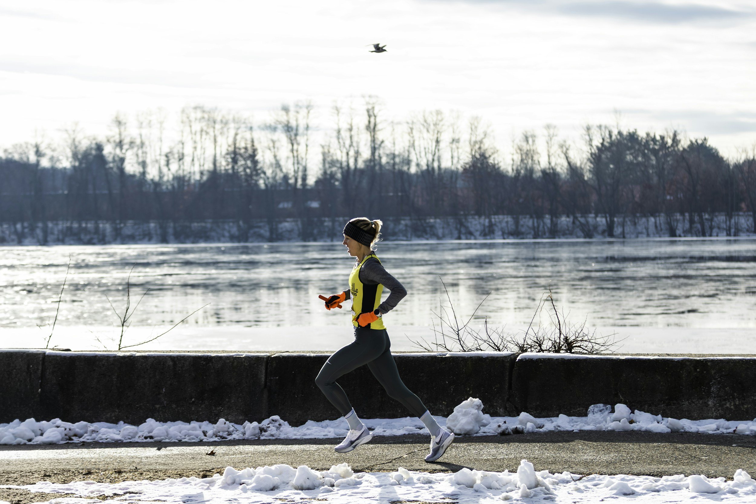 A woman jogs on a snow-covered pavement beside a frozen river during winter, wearing a yellow and gray athletic outfit, gloves, and a headband, with bare trees and a bird flying in the sky in the background.