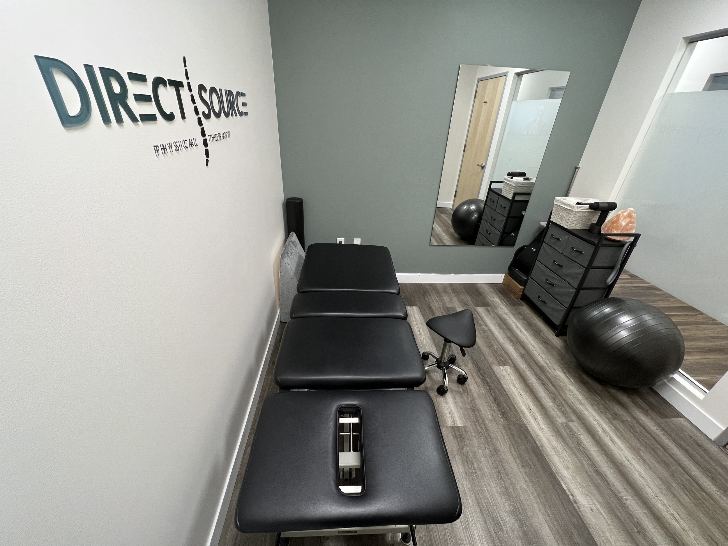 Physical therapy treatment room with black examination table, a small stool, large exercise ball, mirror, and storage drawers.