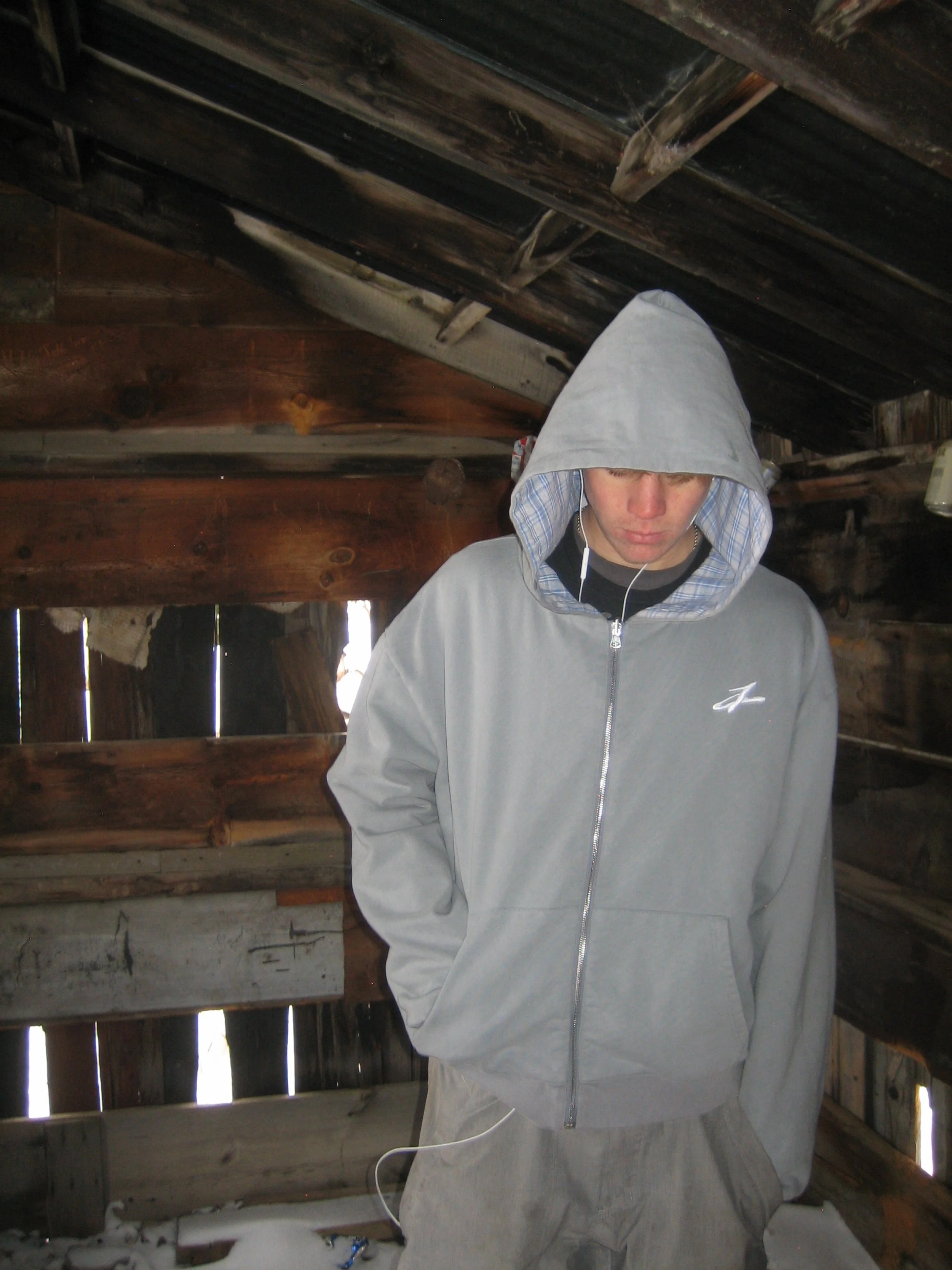 A young man with a hoodie pulled over his head, looking down, inside a wooden barn with weathered walls and visible light coming through gaps in the wood.