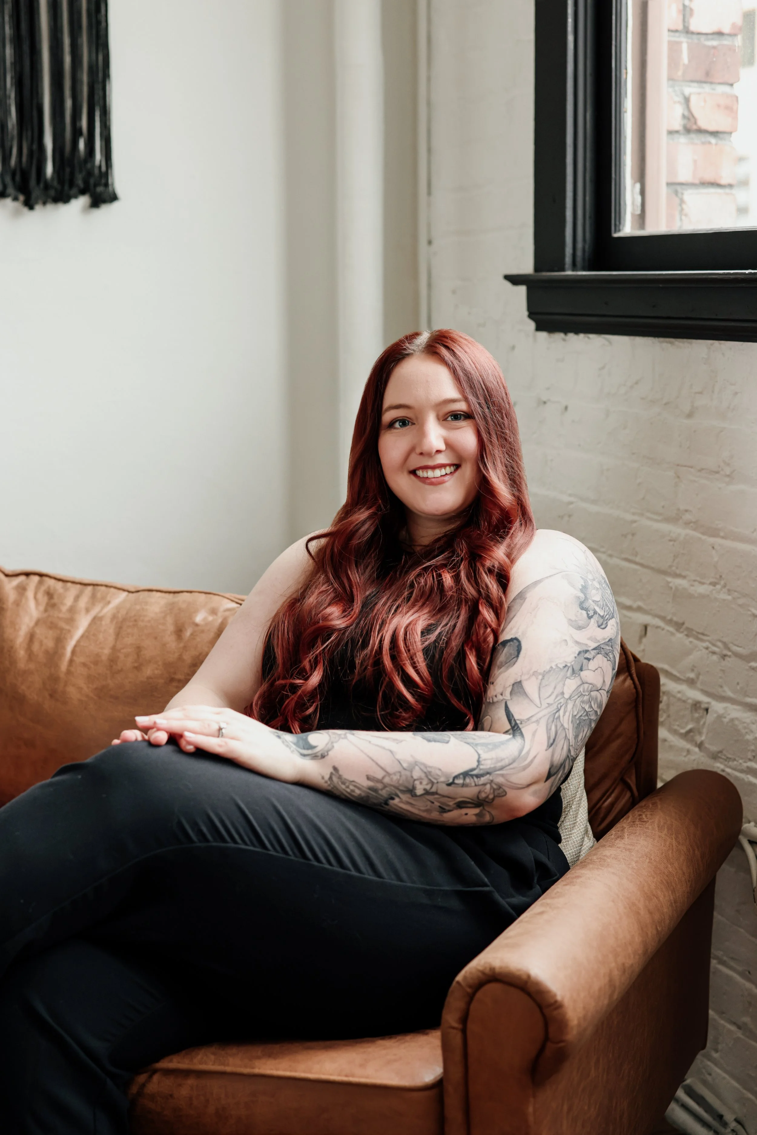 Smiling woman with long red hair, tattoos on her right arm, sitting on a brown sofa in a room with white brick walls and a window.