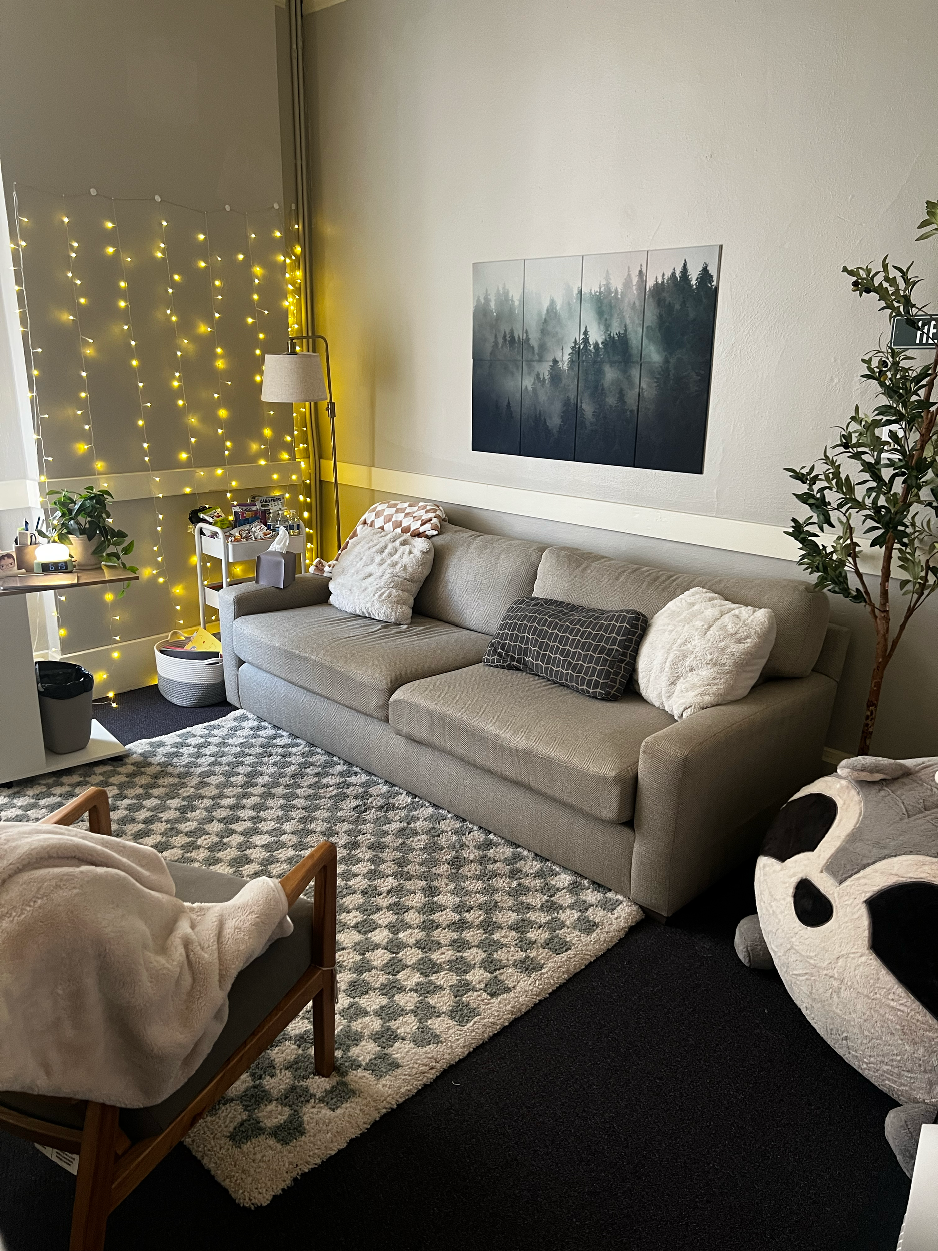 Cozy living room with a beige couch, dinosaur plush toy, patterned pillows, and a green plant next to a wall with a brick-like design. There's a mirror and a soft chair on the side, and a textured rug on the floor.