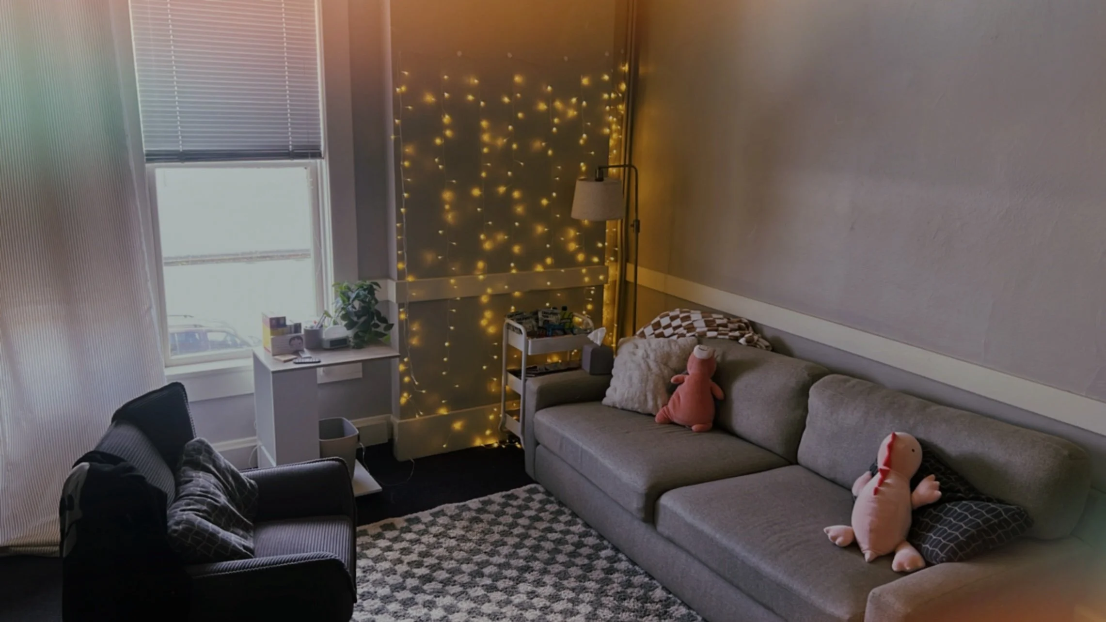 Cozy living room with a beige couch, dinosaur plush toy, patterned pillows, and a green plant next to a wall with a brick-like design. There's a mirror and a soft chair on the side, and a textured rug on the floor.