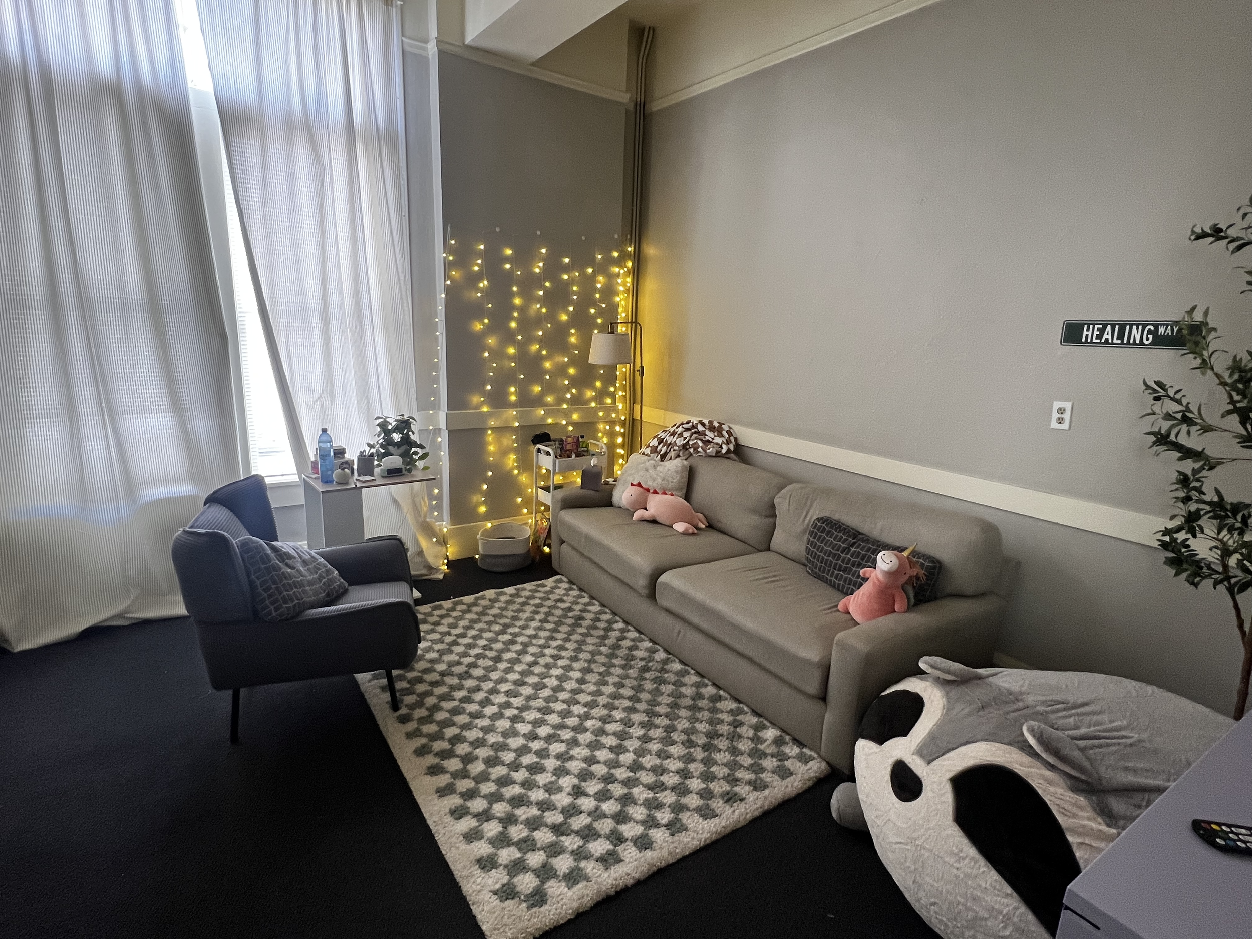 Cozy living room with a beige couch, dinosaur plush toy, patterned pillows, and a green plant next to a wall with a brick-like design. There's a mirror and a soft chair on the side, and a textured rug on the floor.