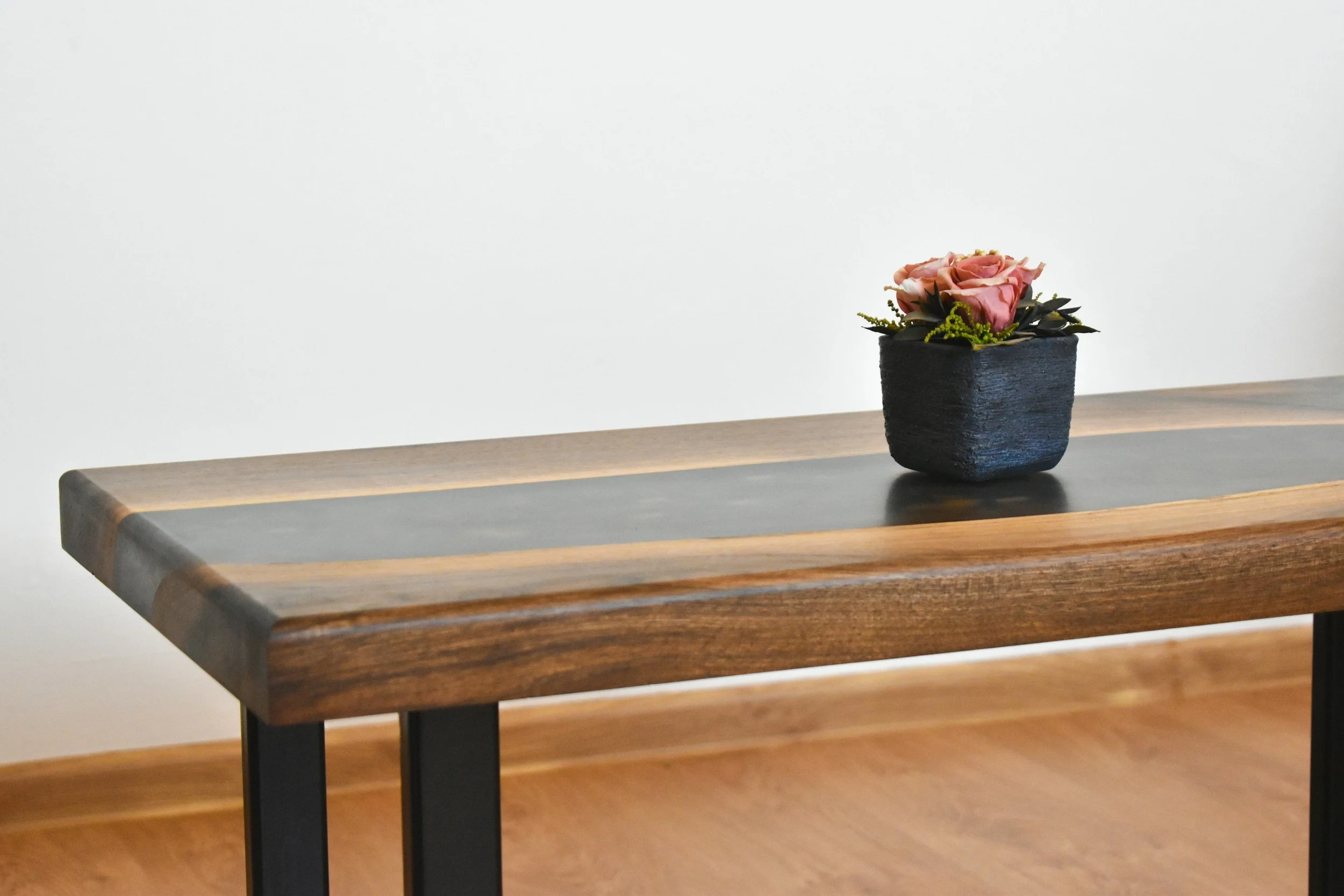 A wooden table with black metal legs and a dark runner in the center, with a small black square pot containing pink roses and greenery on top.