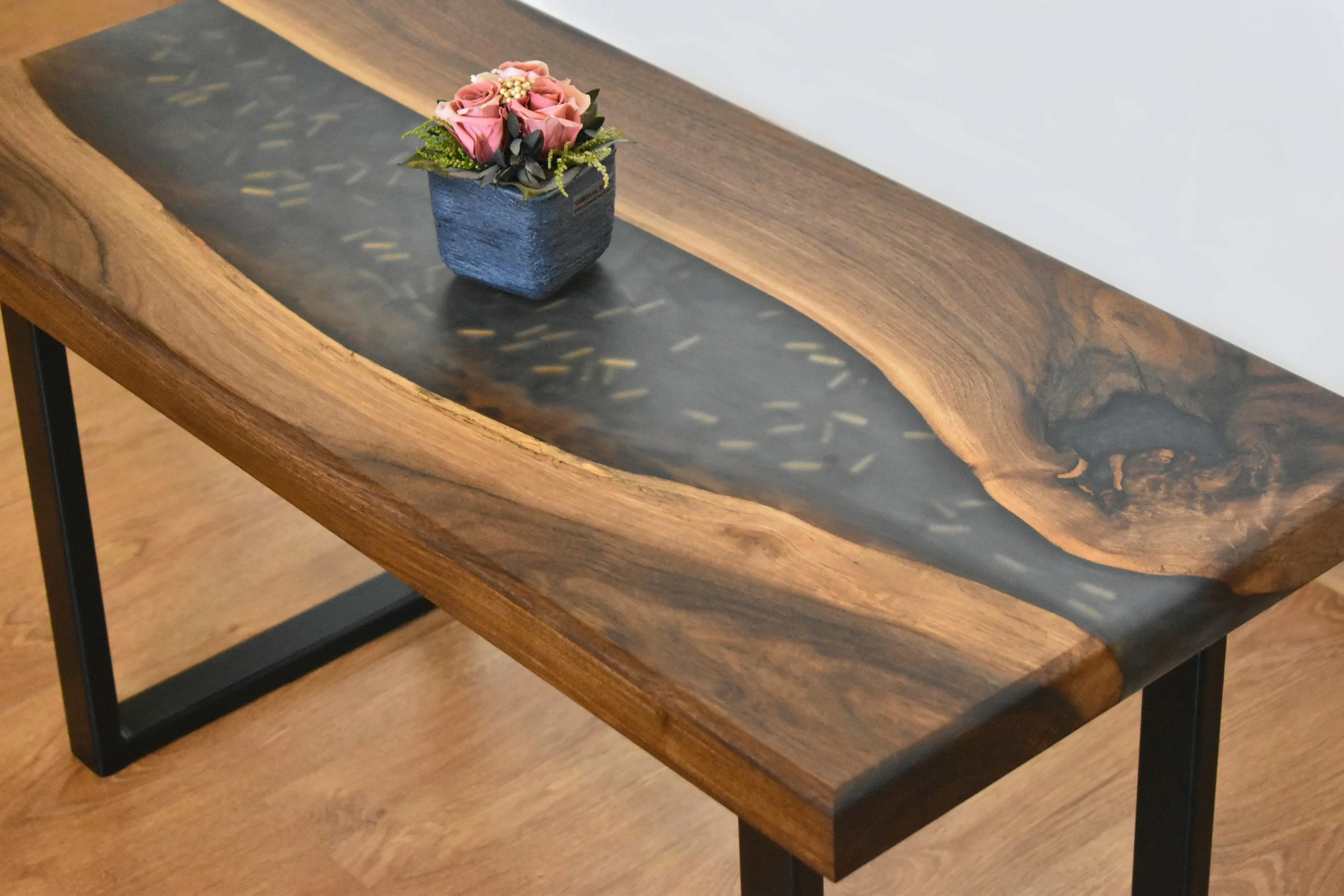 A wooden table with a black resin inlay running through the center and a small pot of pink roses on top.