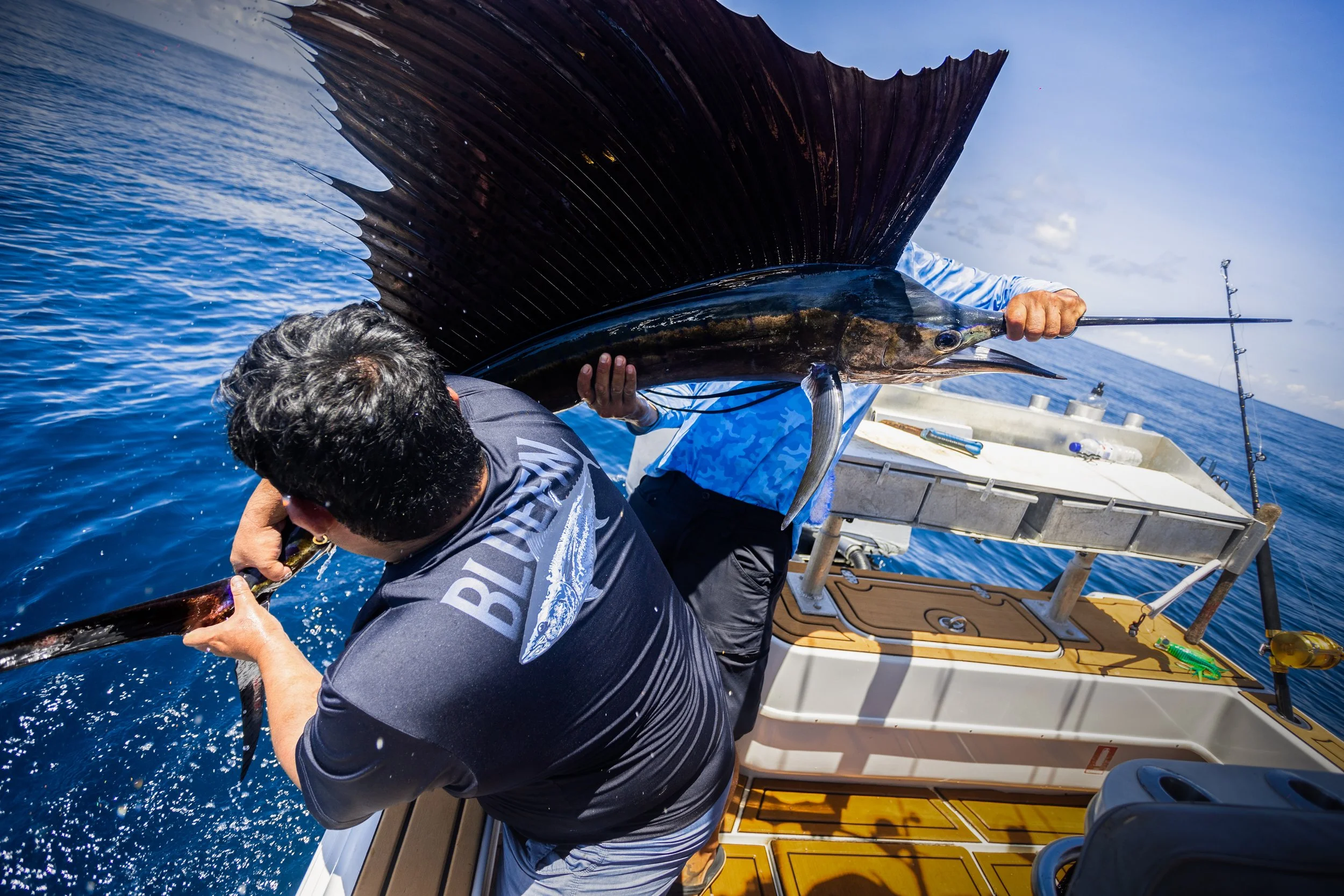 Two men fishing on a boat in the ocean, holding a large fish with dark fins and a long snout.