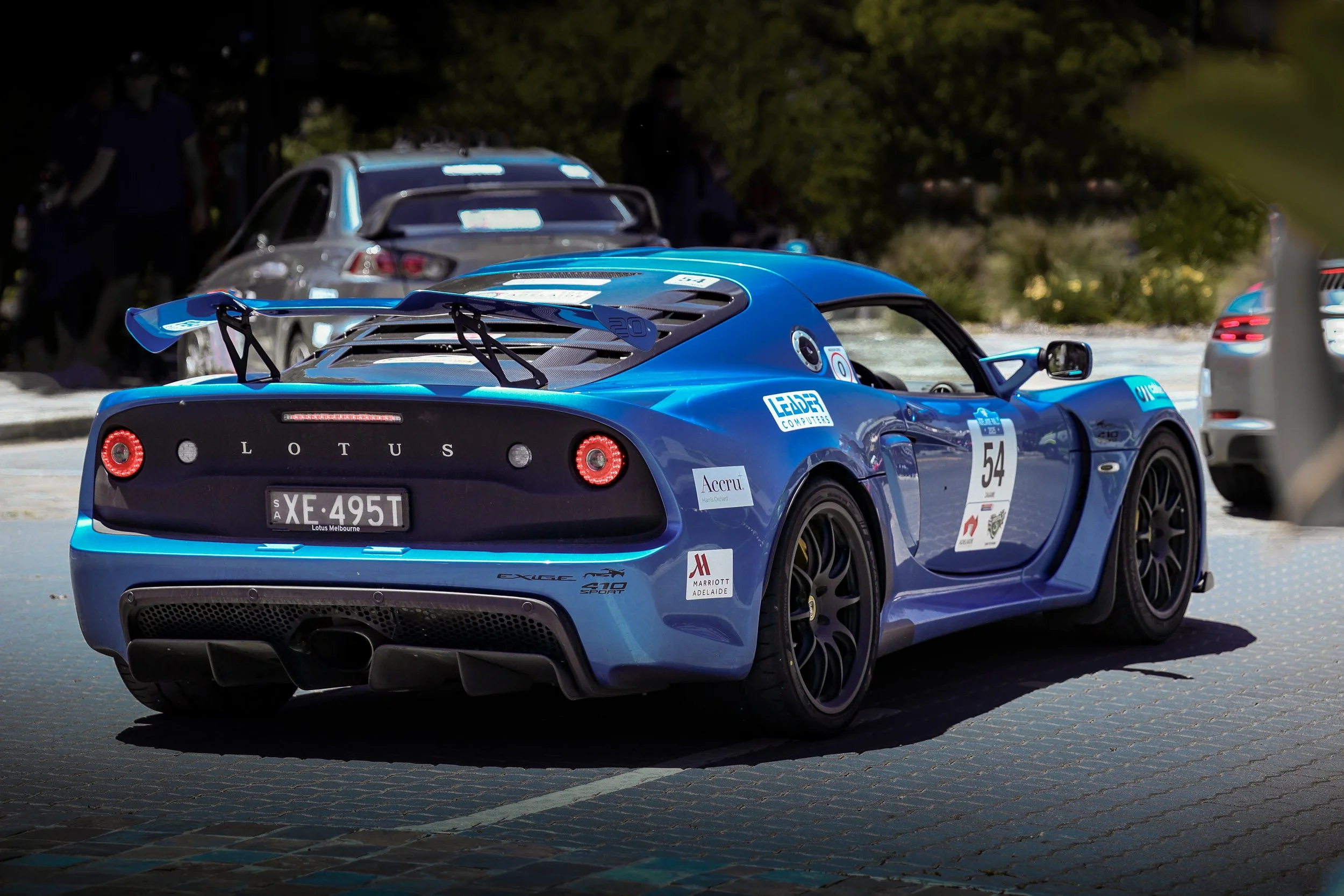 Blue Lotus Exige sports car with race number 54 parked outdoors, surrounded by other cars and trees.