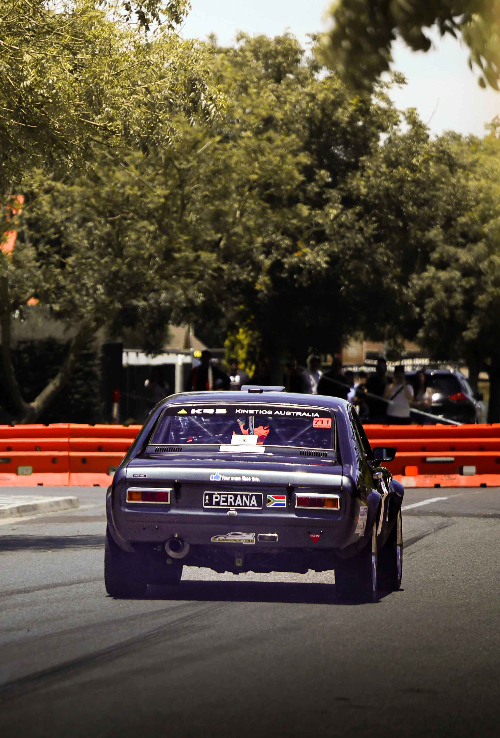 A vintage race car on a city street, with trees and spectators in the background.