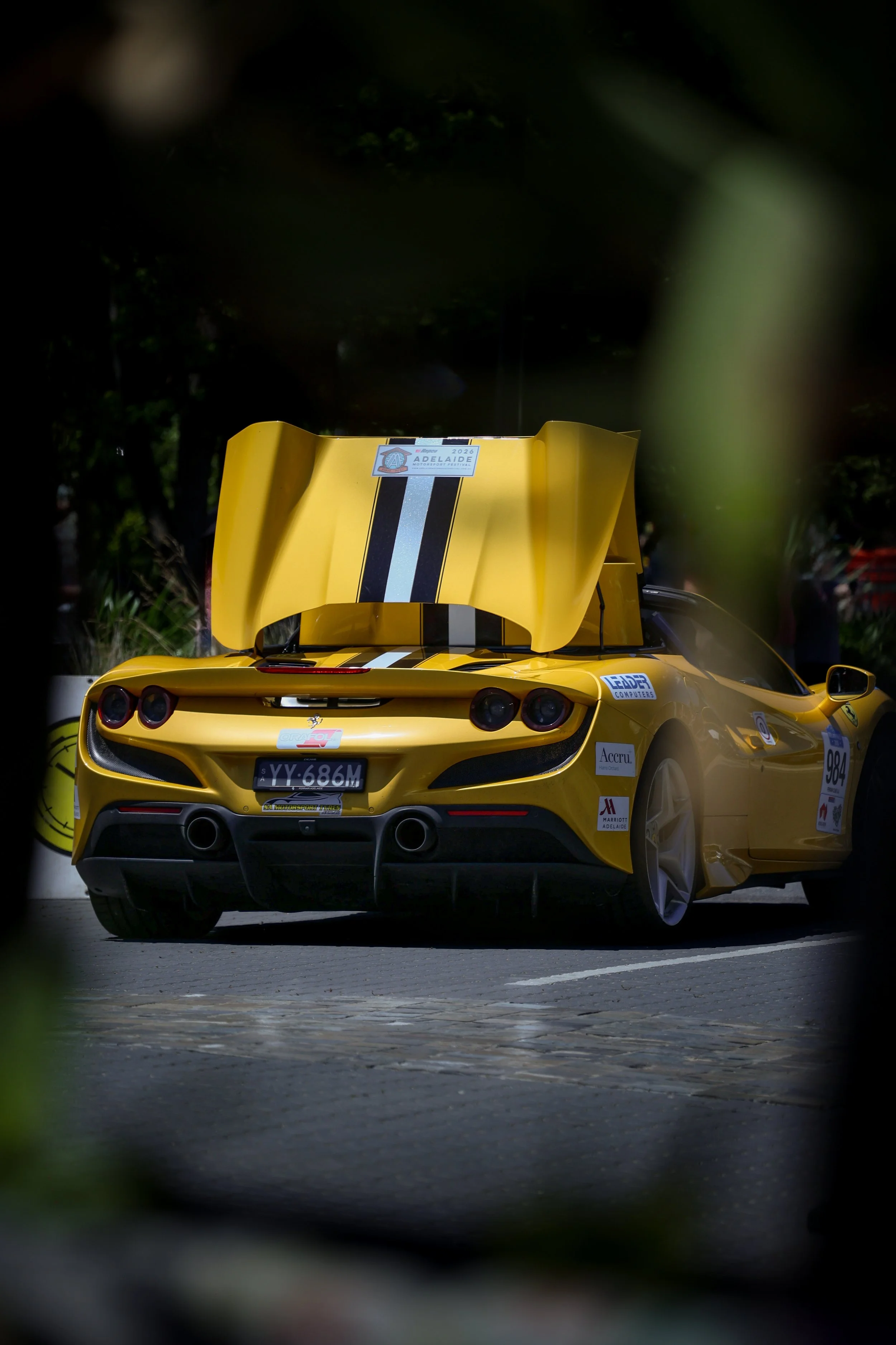 A yellow race car with the hood open, parked outdoors, with racing stickers and the license plate 'YY-686M' visible, surrounded by greenery.