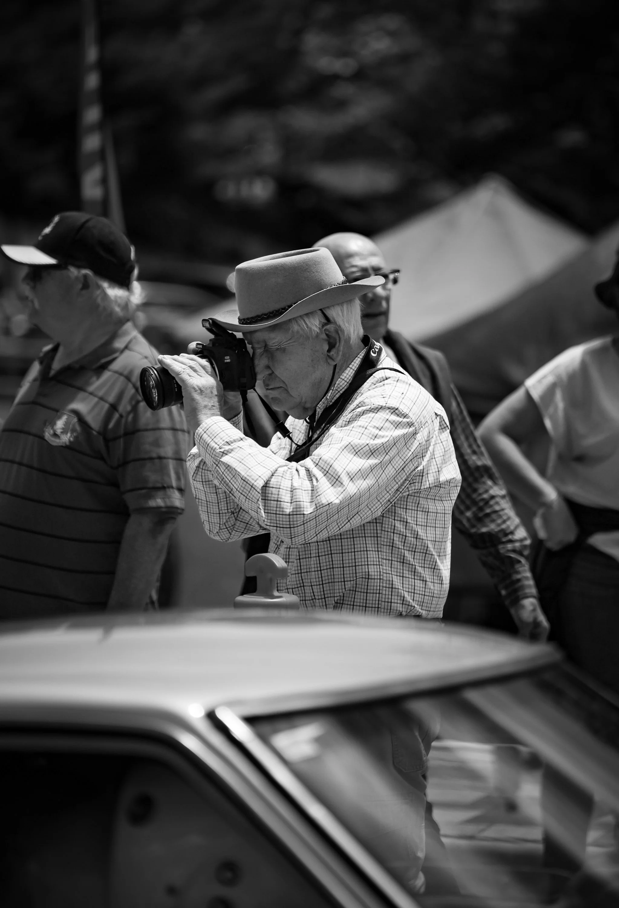 An older man wearing a cowboy hat takes a photograph with a camera at an outdoor event, with other people and tents visible in the background.