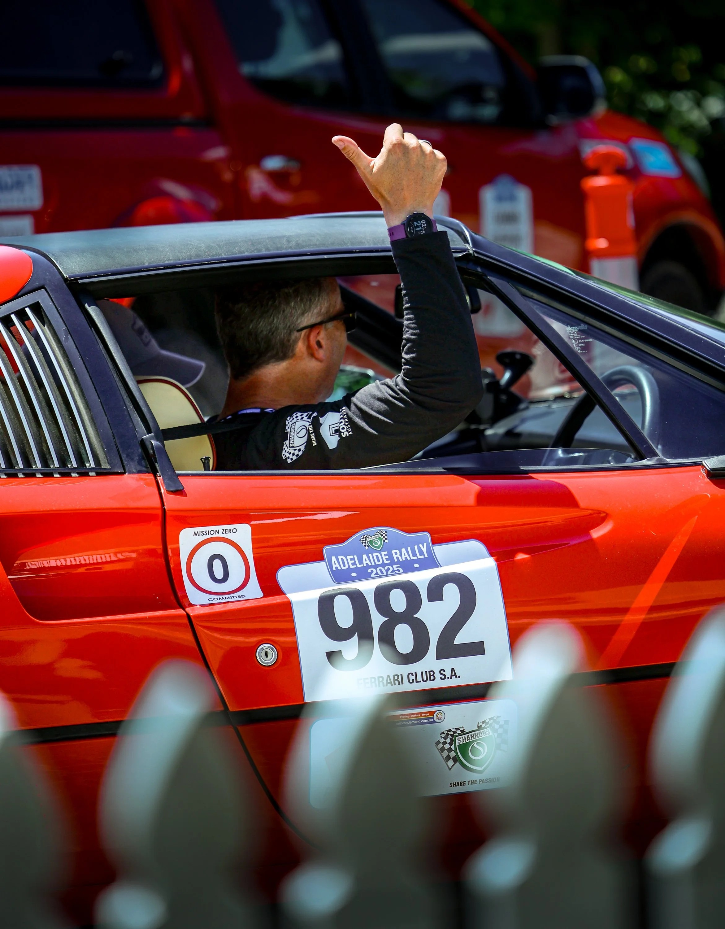A person driving a red race car with the number 982 and an Adelaide Rally 2025 sticker, giving a thumbs up through the open window.