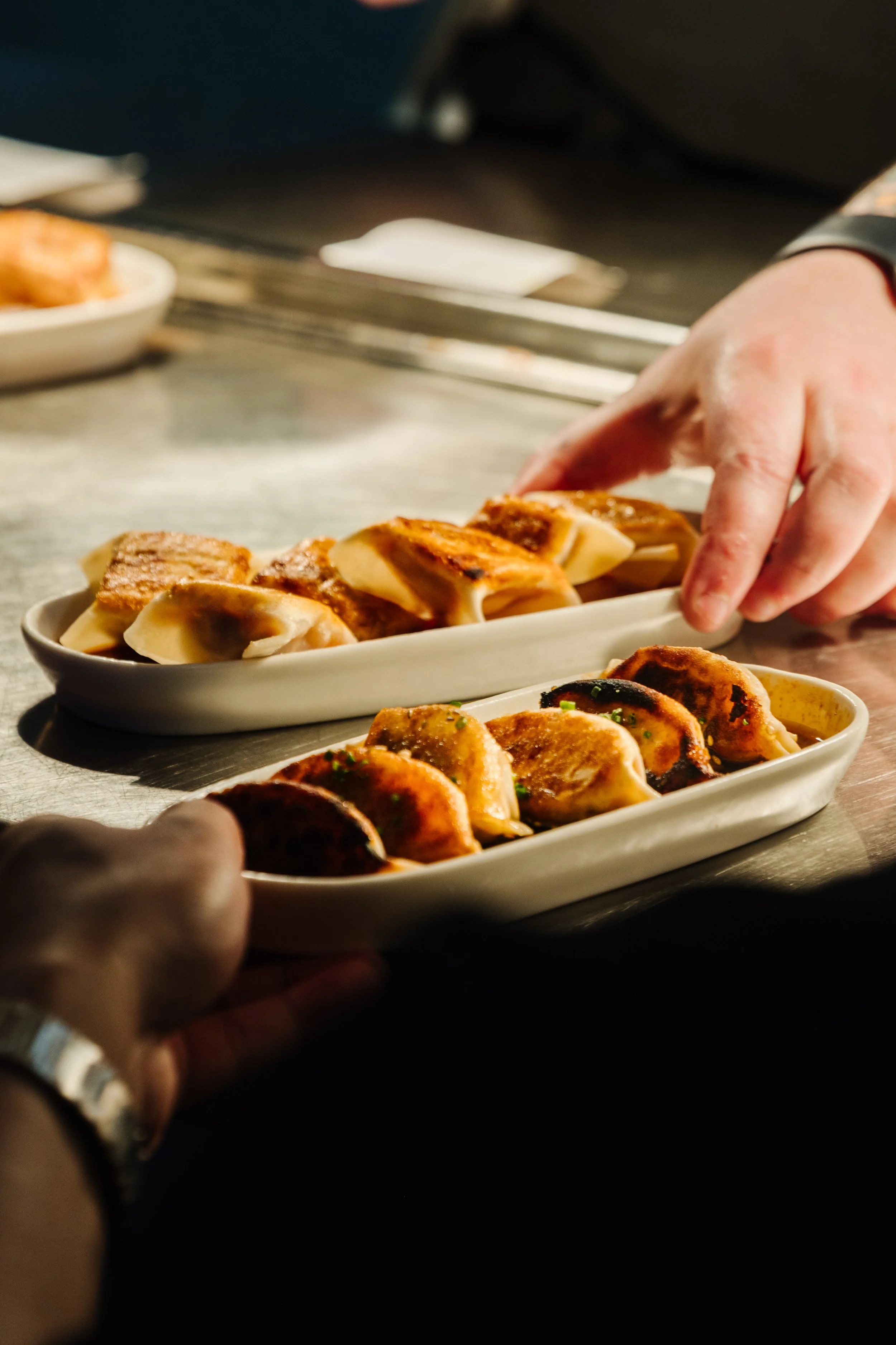 Hands passing trays of cooked Asian dumplings across a stainless steel countertop.