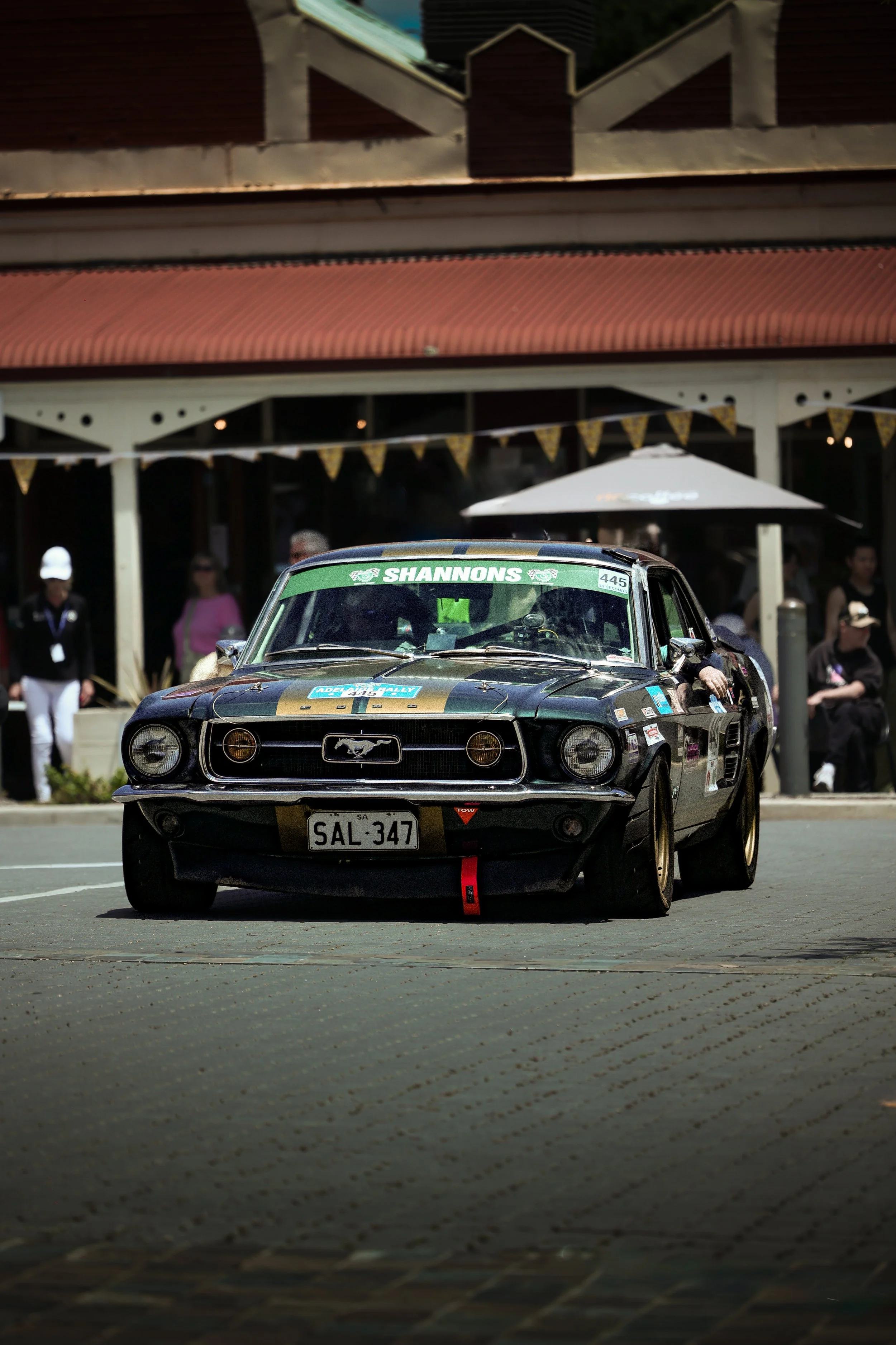 A black vintage Ford Mustang race car with gold accents on a street in front of a building with a red roof and people watching in the background.