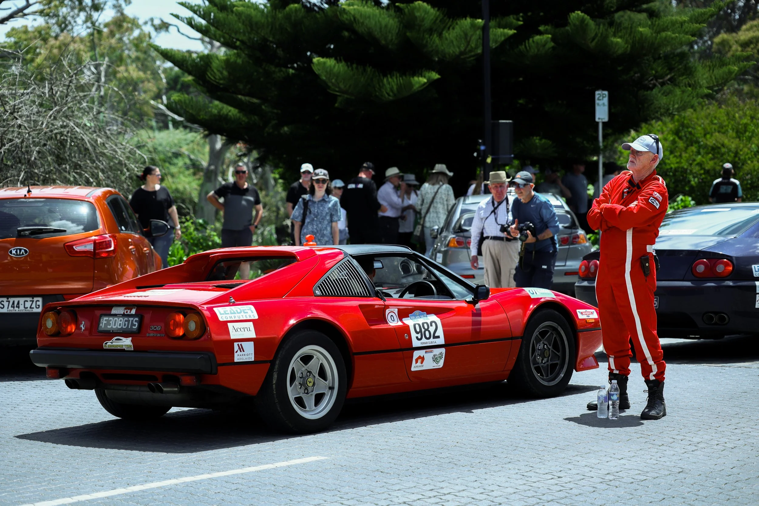 Red classic race car with the number 982, surrounded by spectators and a man in a red racing suit standing with arms crossed and water bottles at his feet, in a parking lot with green trees in the background.