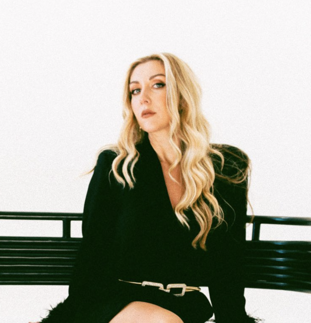 Woman with long blonde hair wearing a black outfit, sitting on a bench against a plain background.
