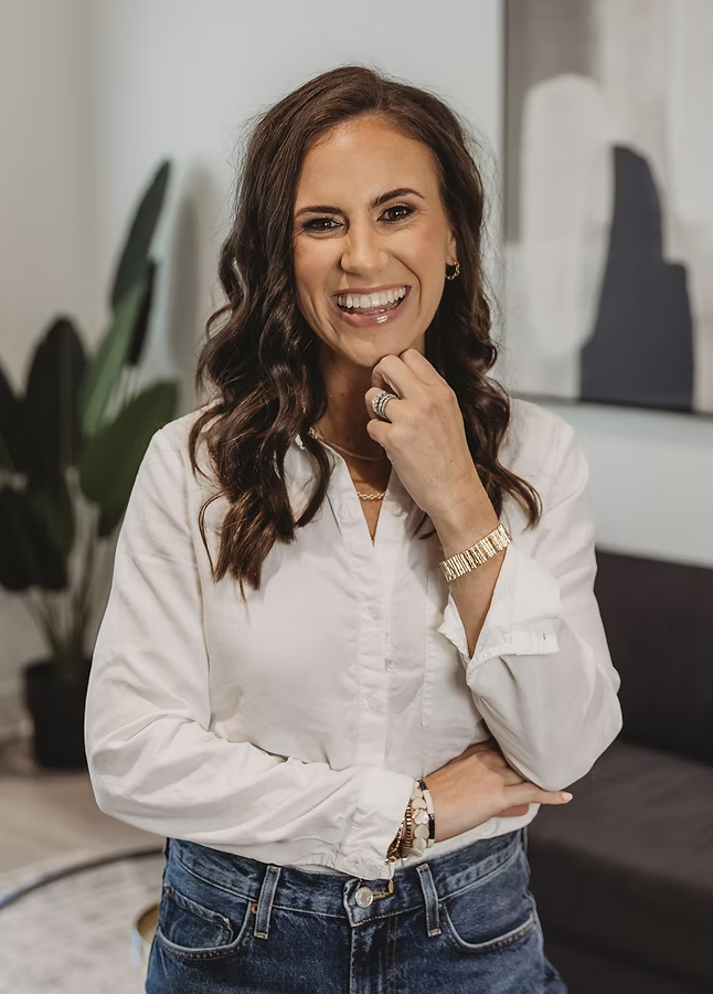 Smiling woman in white blouse and jeans with hand on chin.