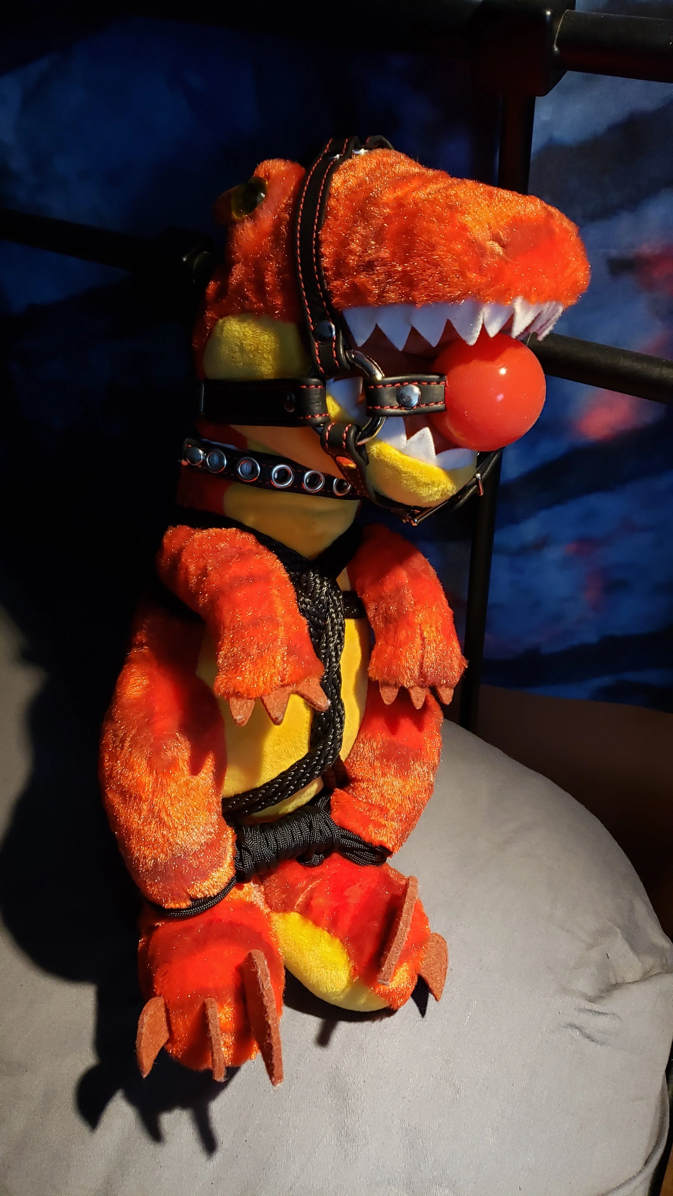 A plush toy of a dinosaur wearing a teddy bear costume with a clown nose, collar, and harness, sitting on a surface with a dark background.