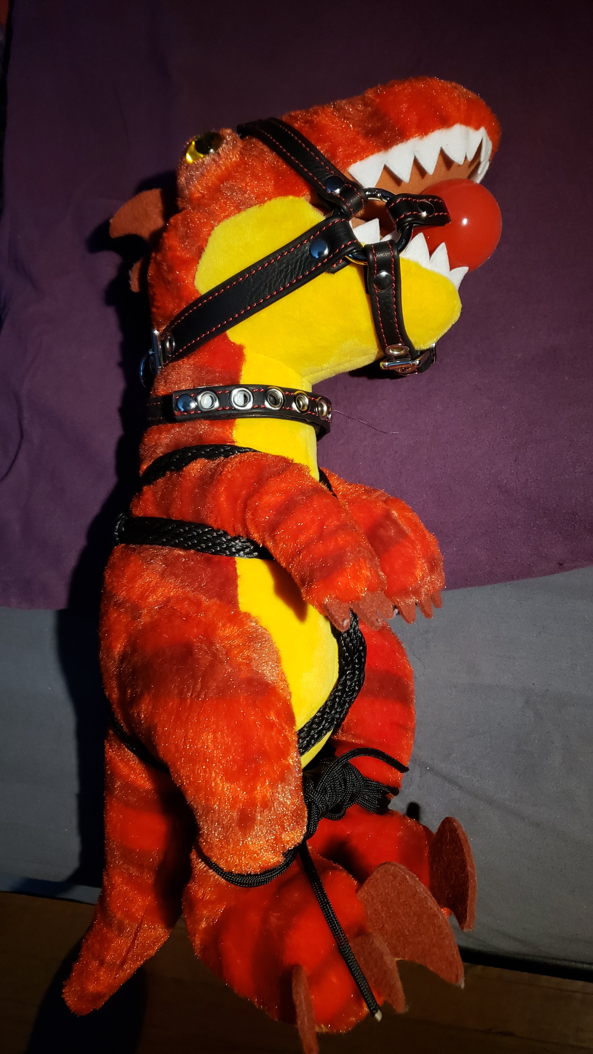 A plush dinosaur toy dressed in a harness with black straps, metal studs, and laces. The toy is red and yellow with a red ball in its mouth, placed on a dark surface with a purple background.