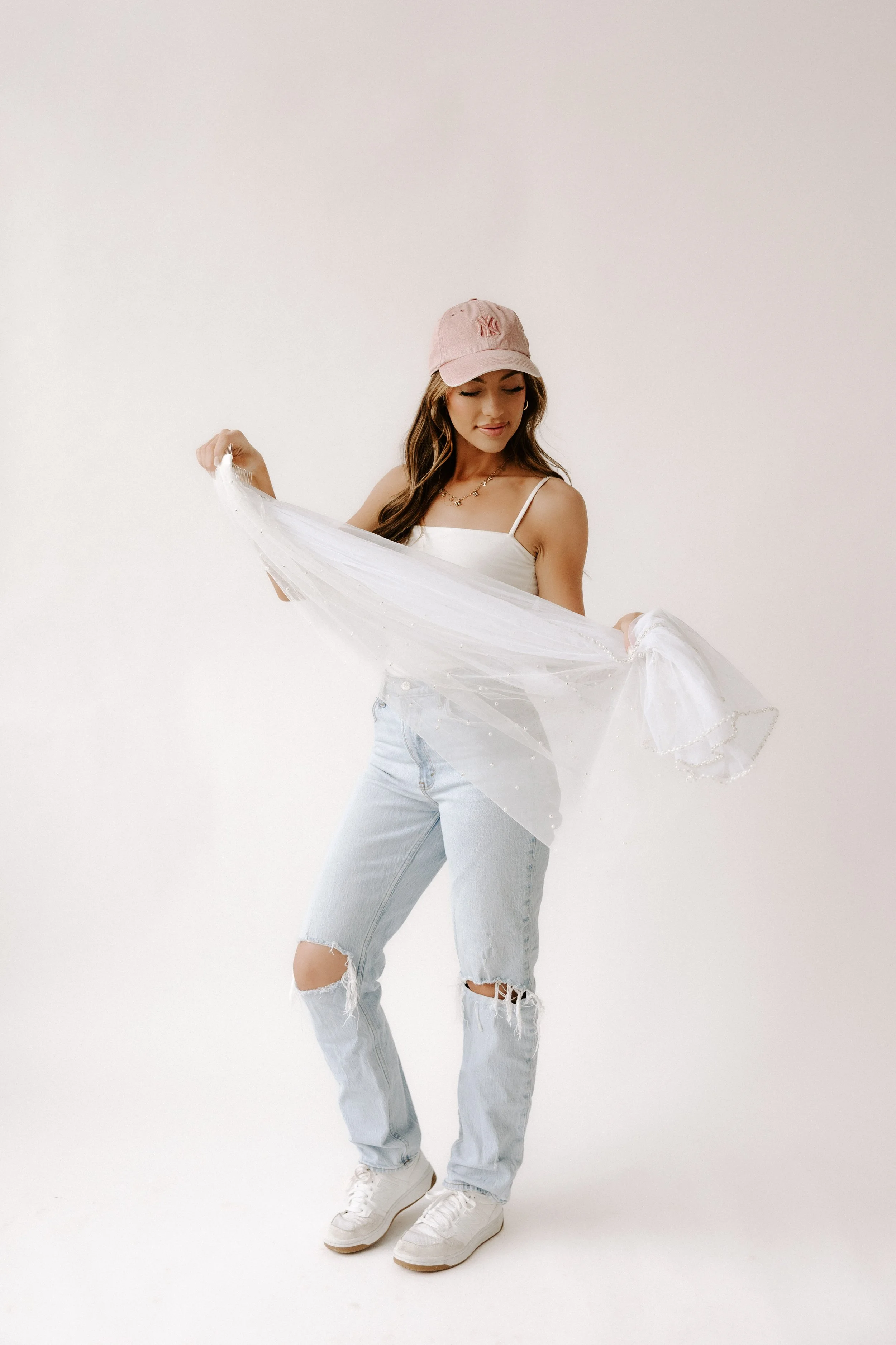 Young woman wearing a pink baseball cap, white tank top, ripped light blue jeans, and white sneakers, holding a sheer, decorated fabric against a plain white background.