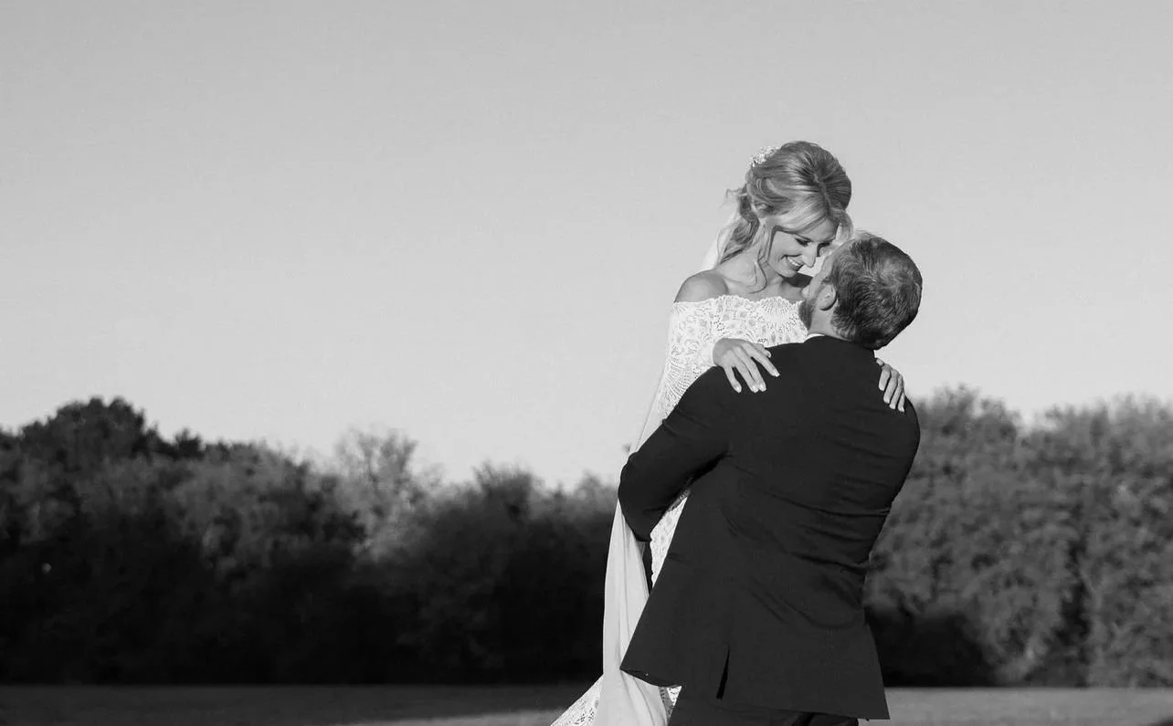 A man lifting a woman in a field with trees in the background, smiling and looking at each other.