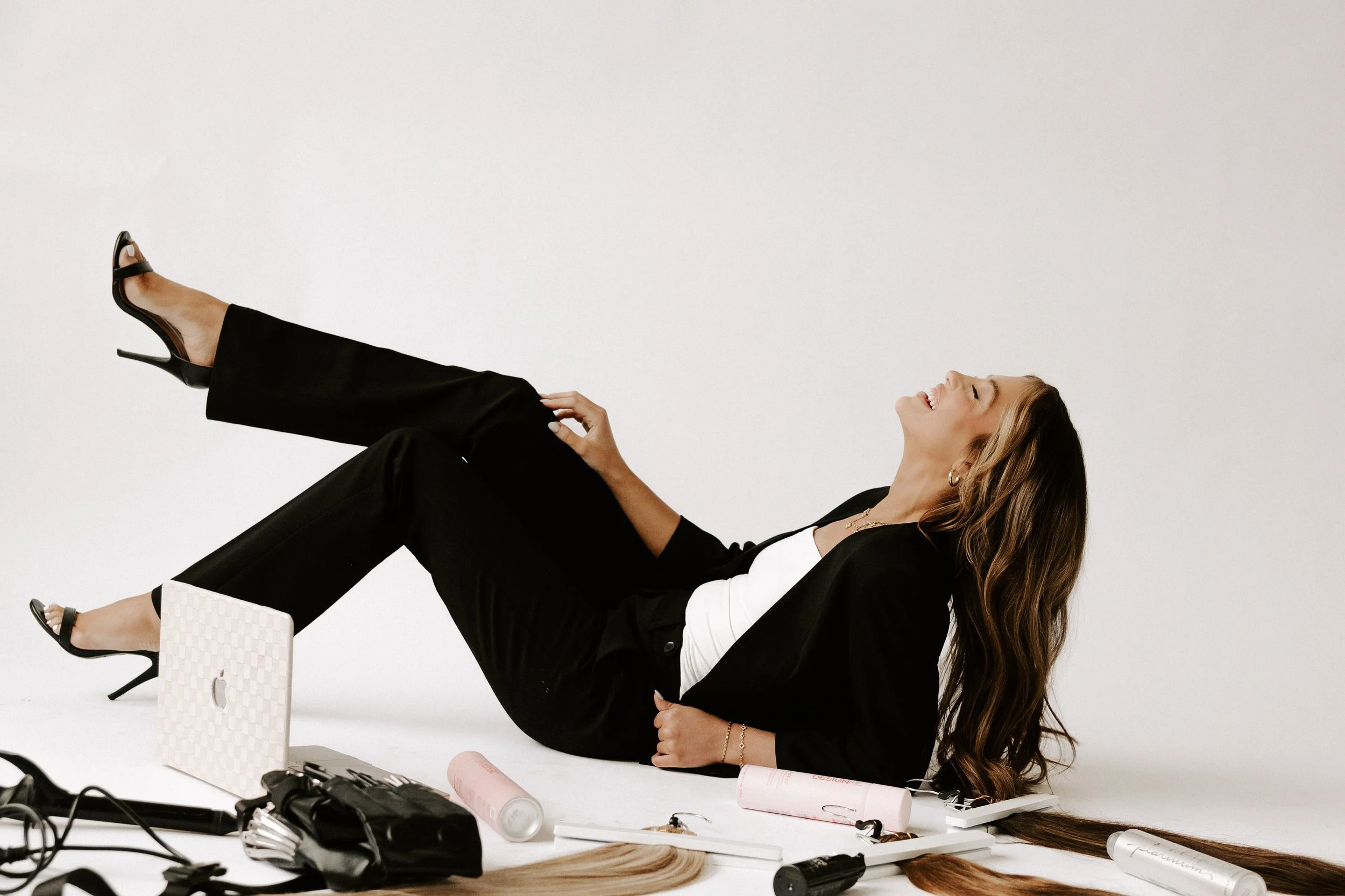 Woman lying on the floor, laughing, surrounded by beauty and hair styling products and tools.