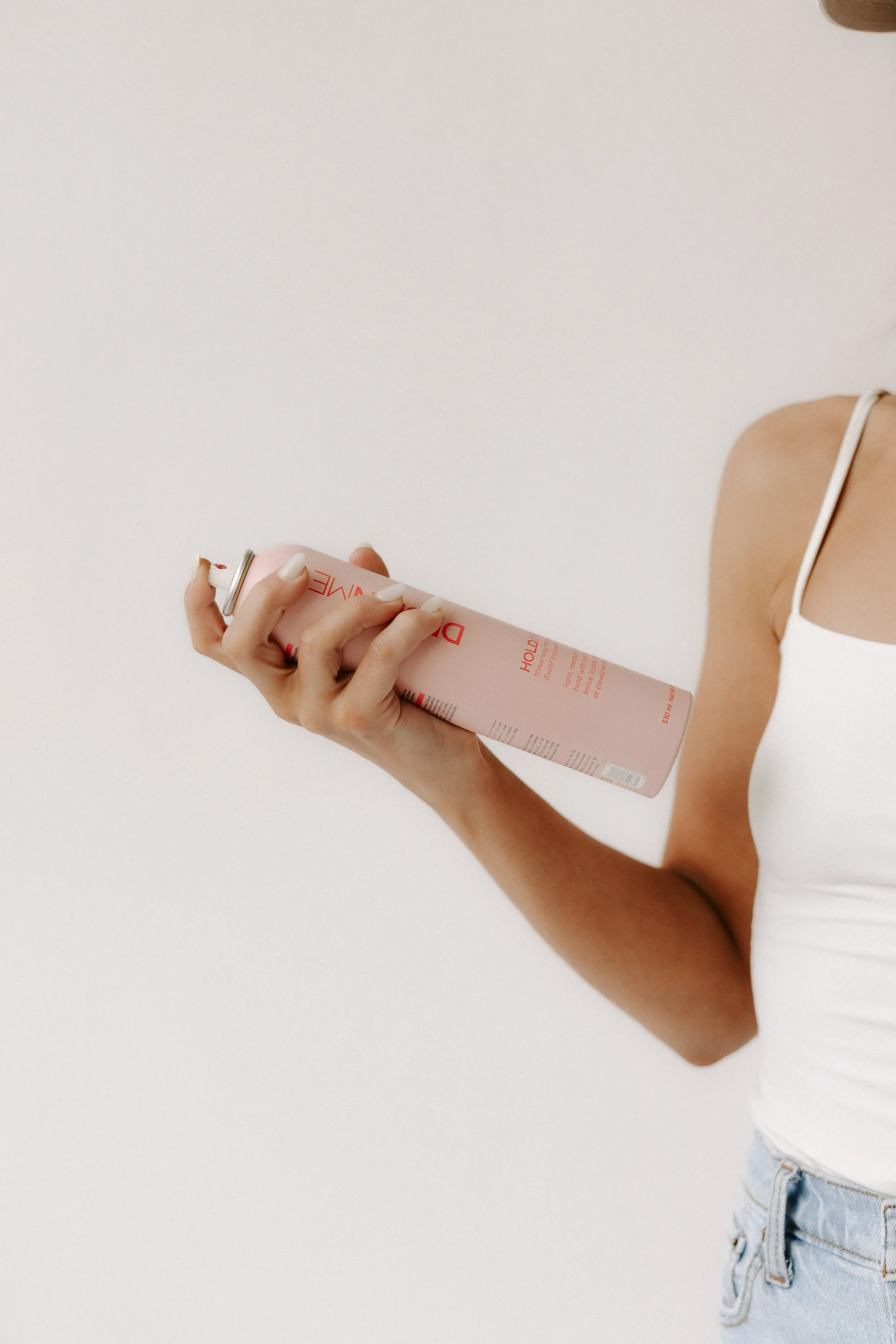 A woman wearing a white tank top and light blue jeans is holding a pink can of dry shampoo with her right hand, against a plain white background.