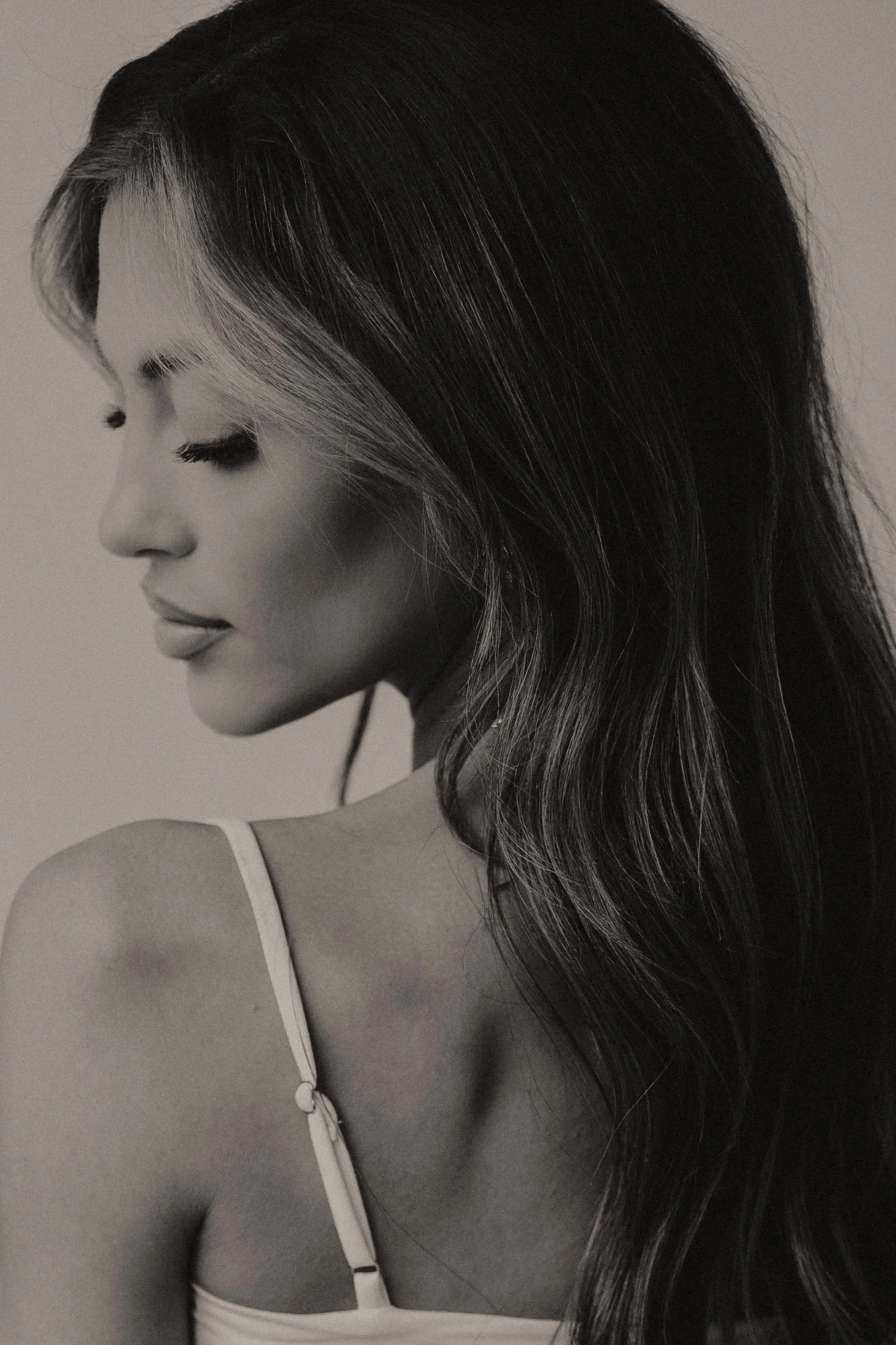 Black and white portrait of a woman with long wavy hair, her eyes closed and head slightly tilted downward, wearing a thin strap top.