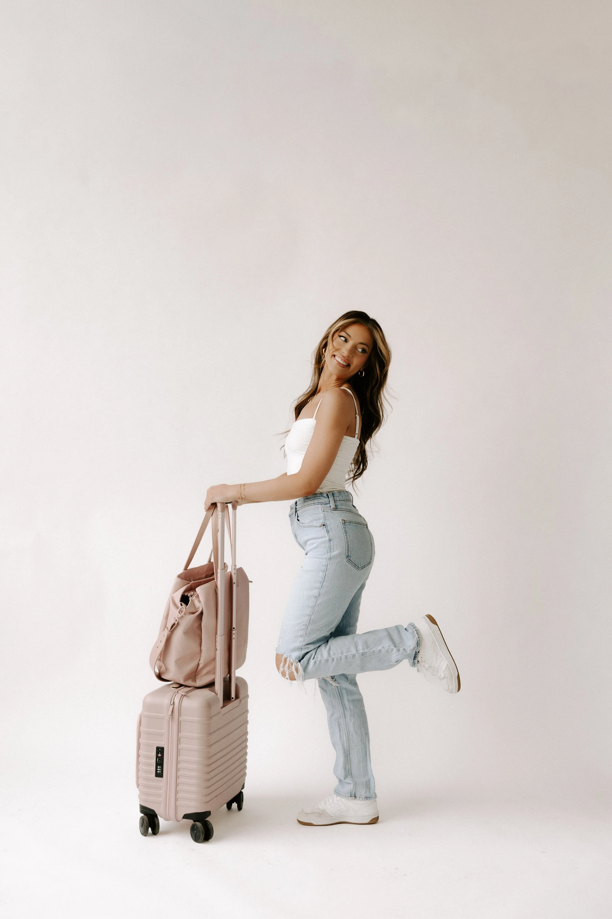 A woman with long wavy hair wearing a white tank top, light blue distressed jeans, and white sneakers, holding a pink rolling suitcase and a pink tote bag, standing in a studio with a plain light background, smiling and lifting one leg.