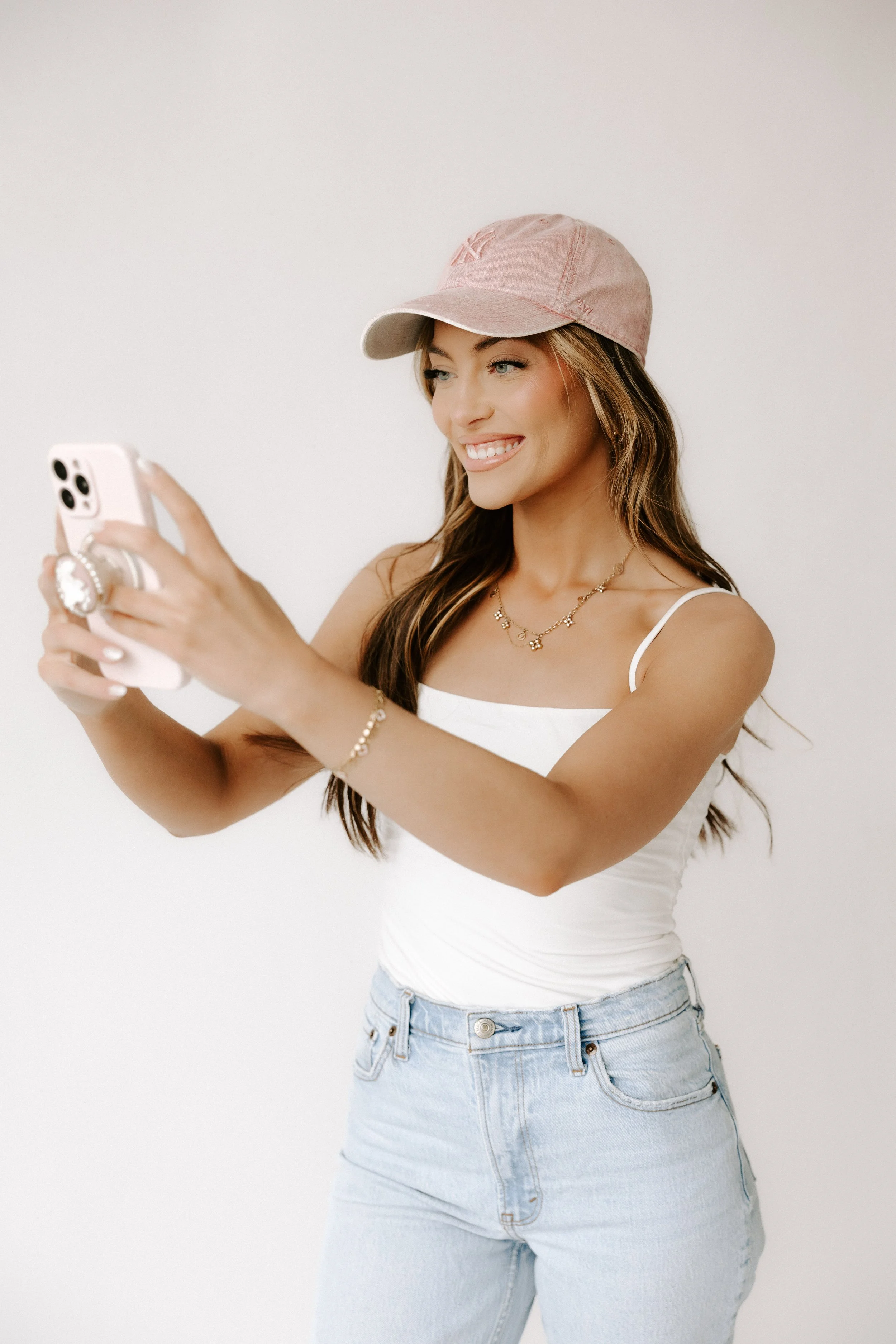A woman with long brown hair, wearing a pink baseball cap, white tank top, and light blue jeans, takes a selfie against a plain white wall.